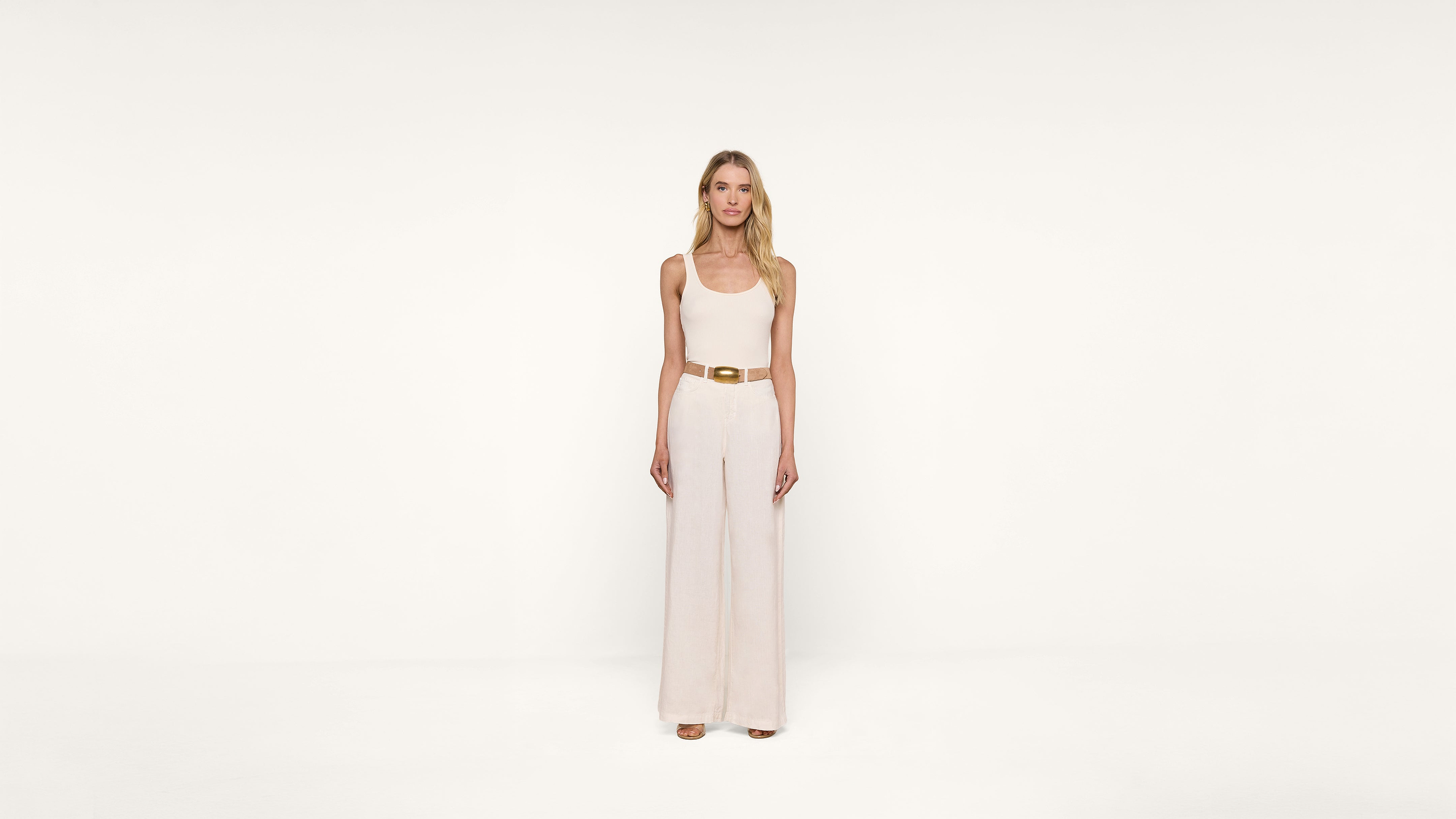 A woman with long blonde hair stands facing forward, wearing a sleeveless beige jumpsuit with wide legs and a tan belt, against a plain white background.