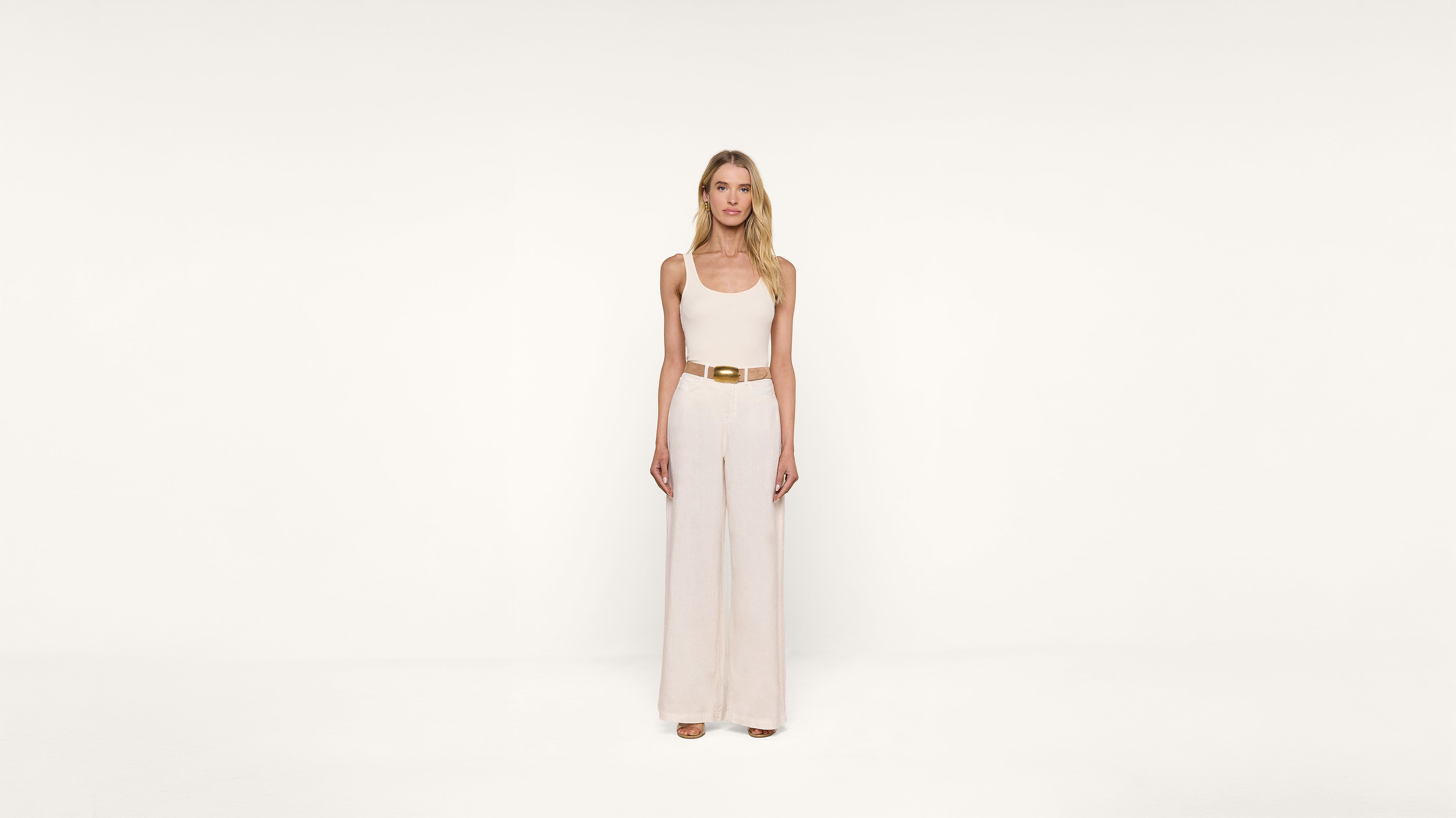 A woman with long blonde hair stands facing forward, wearing a sleeveless beige jumpsuit with wide legs and a tan belt, against a plain white background.