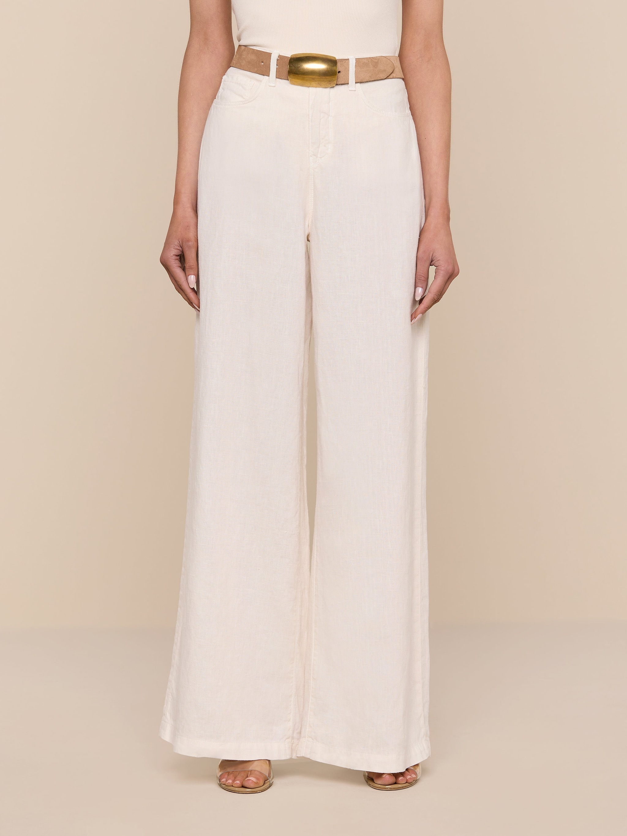Wearing the Amara Linen Wide-Leg Pant in light beige, styled with a matching beige belt and brown open-toed shoes, the person stands against a plain white background with their upper body partially out of frame.
