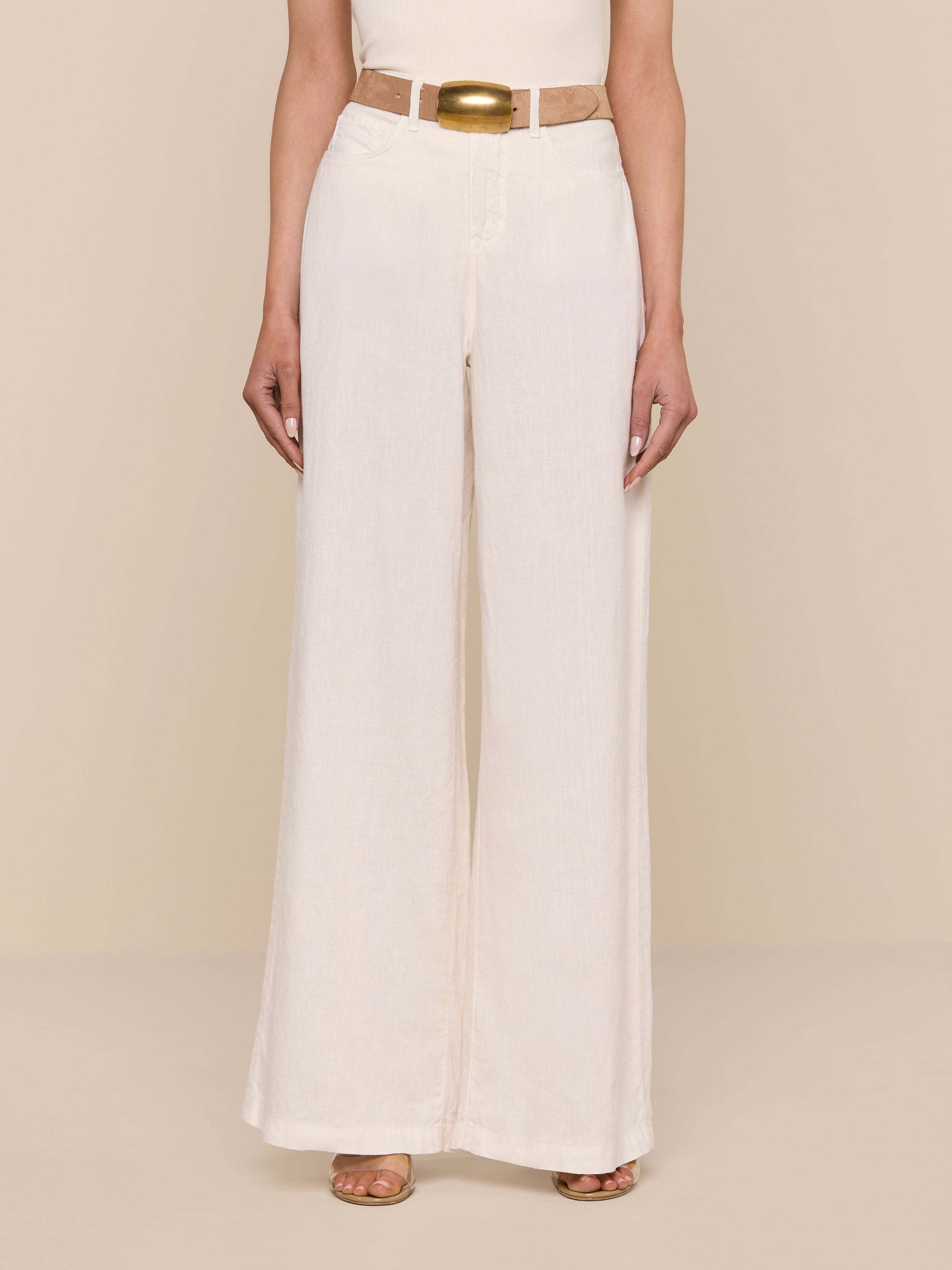Wearing the Amara Linen Wide-Leg Pant in light beige, styled with a matching beige belt and brown open-toed shoes, the person stands against a plain white background with their upper body partially out of frame.