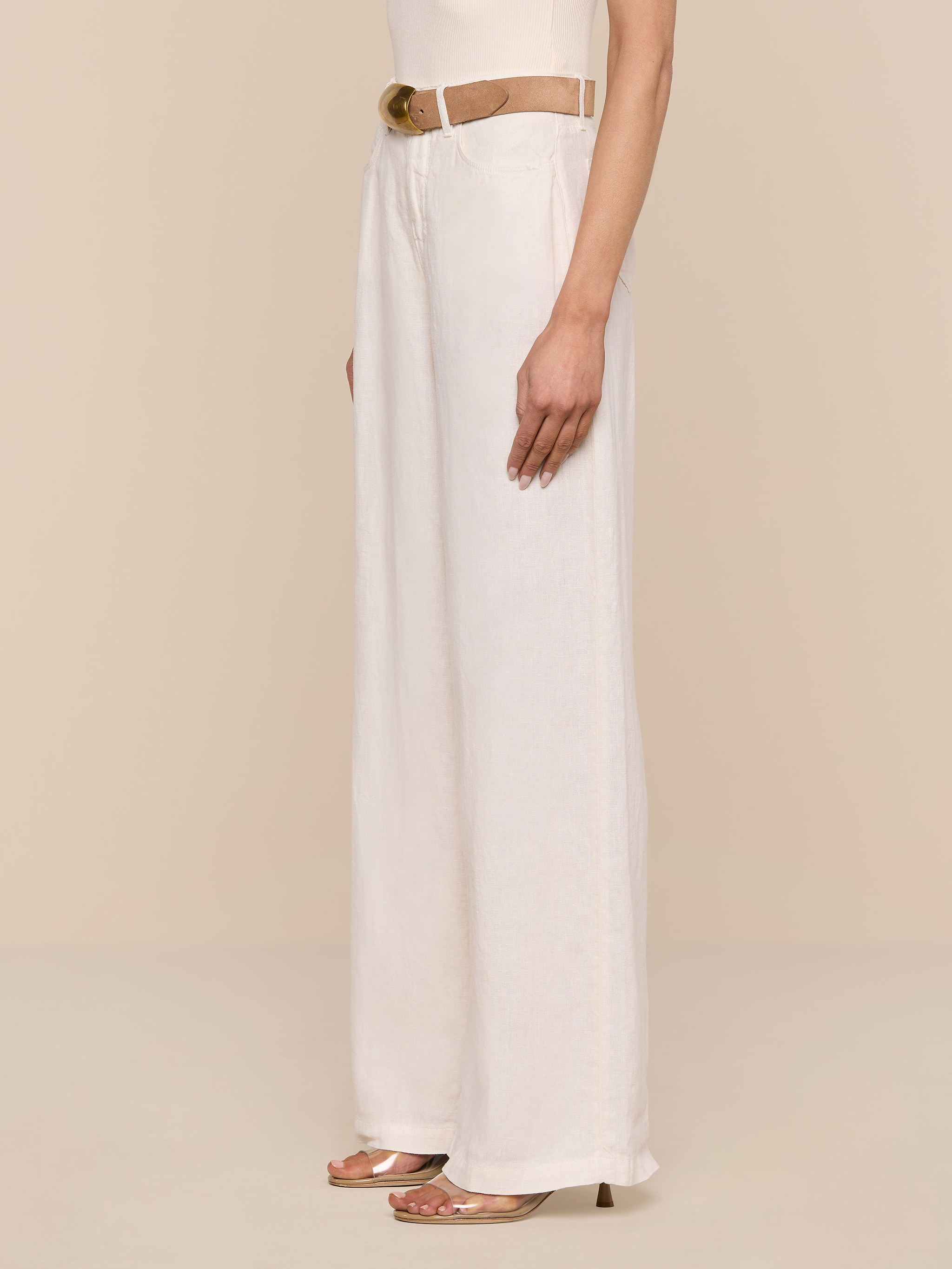 A person wearing the Amara Linen Wide-Leg Pant in cream, styled with a beige belt and open-toe heels, stands against a plain white background, shown from the waist down in a side view.