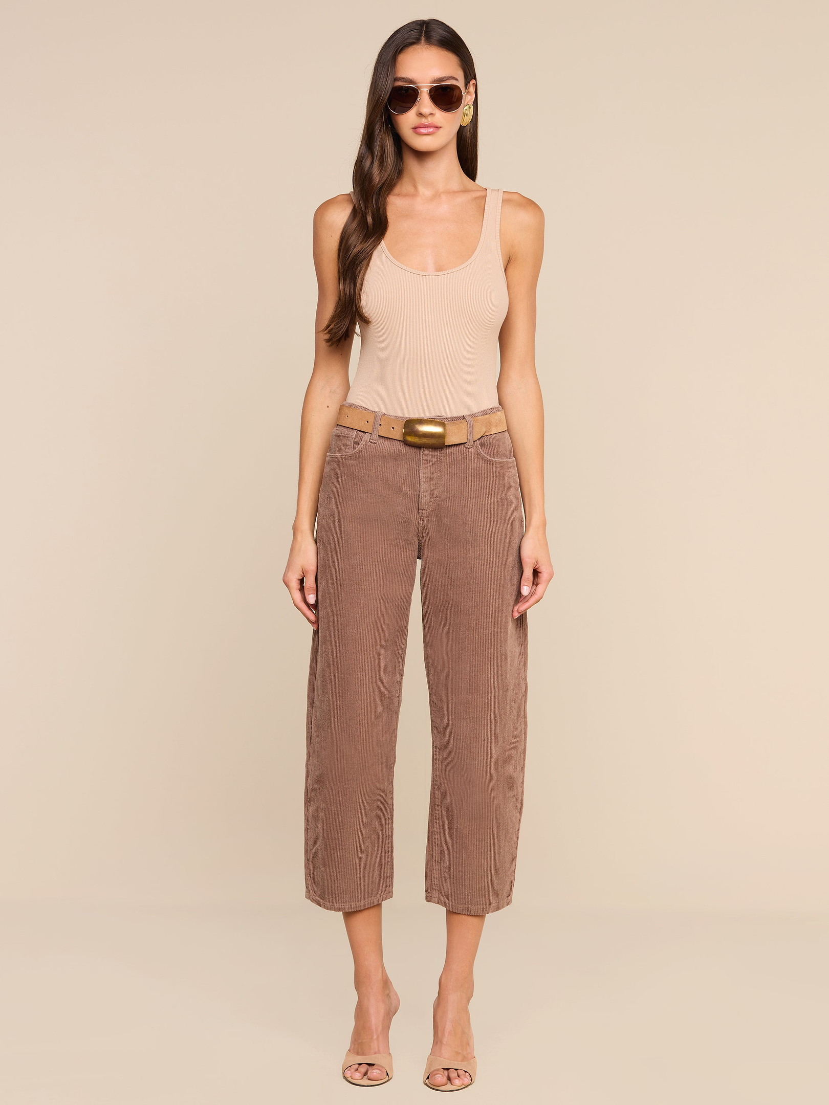 A woman with long hair, wearing sunglasses, a beige tank top, the high-rise brown Mallis Corduroy Bow-Leg Pant, and open-toe heels stands against a plain white background.