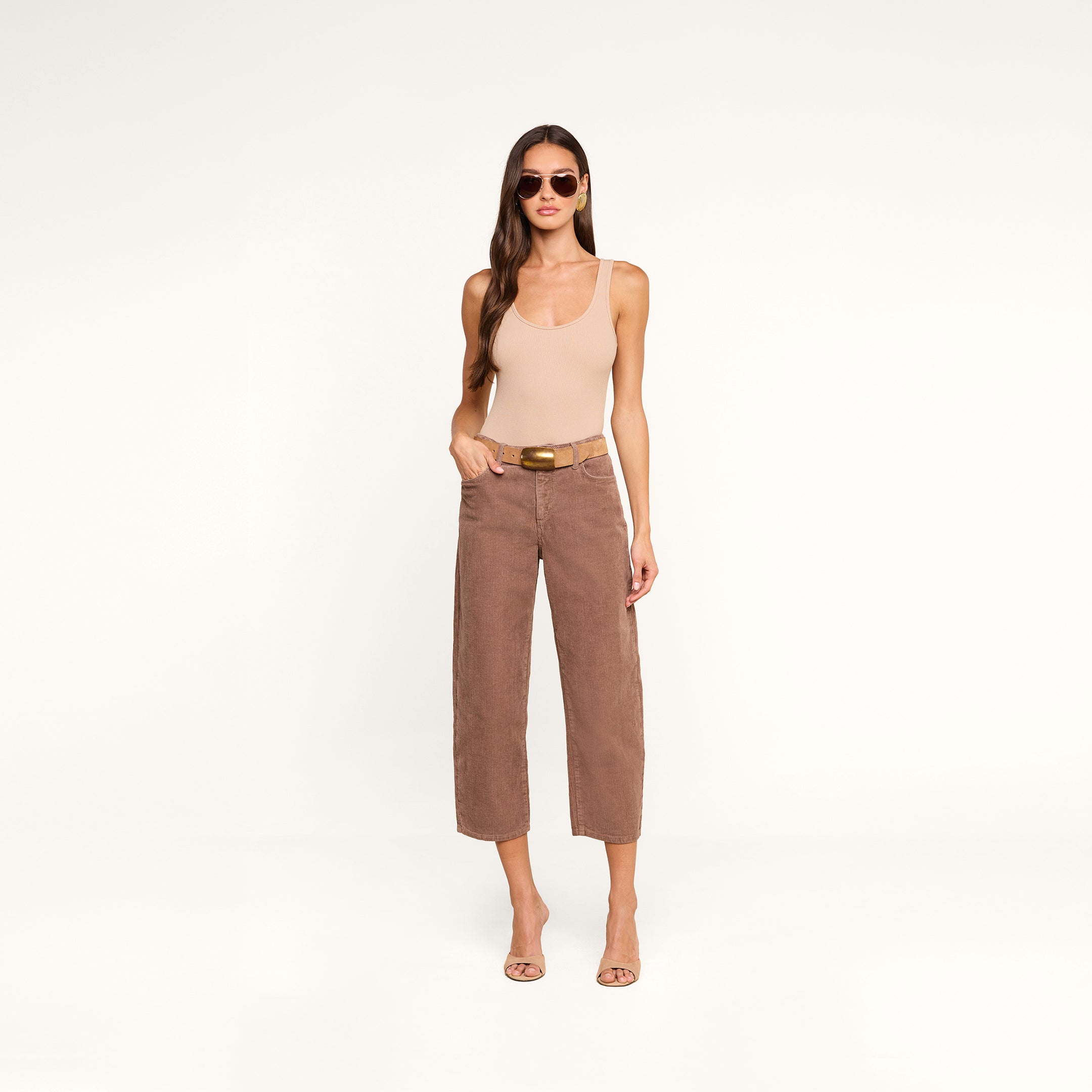 A woman wearing sunglasses, a beige sleeveless top, the Mallis Corduroy Bow-Leg Pant in brown high rise wide-leg style, and heeled sandals stands against a white background with her hands in her pockets.