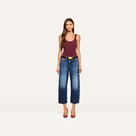 A woman stands facing forward, wearing a maroon tank top, the Mallis Bow-Leg Jean in indigo wash paired with a brown belt, and red heels, against a plain white background.