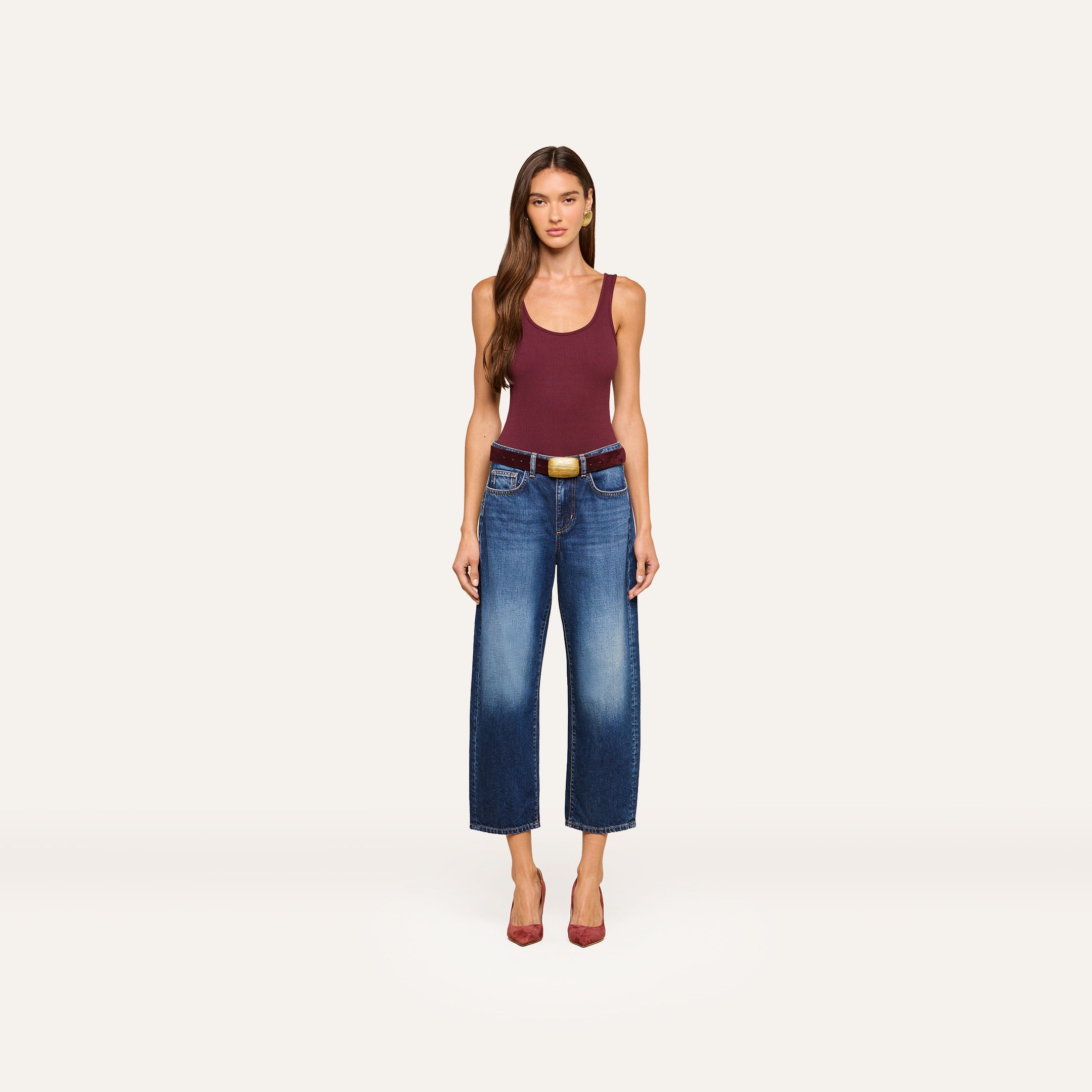 A woman stands facing forward, wearing a maroon tank top, the Mallis Bow-Leg Jean in indigo wash paired with a brown belt, and red heels, against a plain white background.