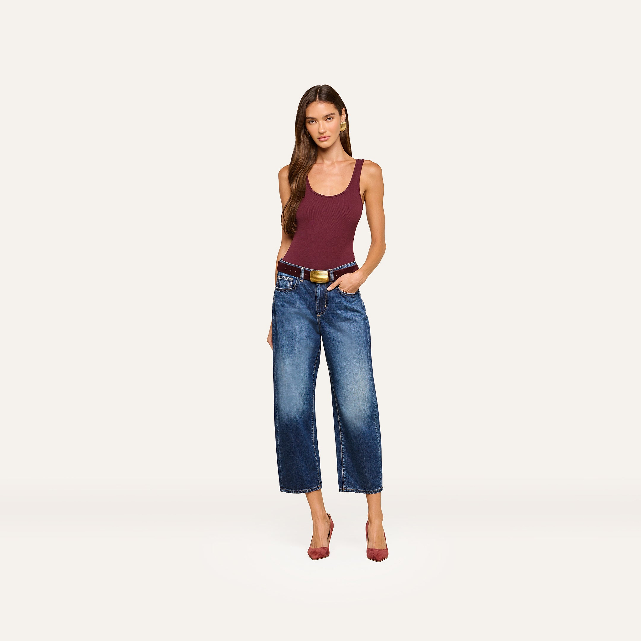 A woman in a burgundy tank top, black belt, and red heels stands against a plain white background, looking at the camera with one hand in her pocket while wearing the Mallis Bow-Leg Jean in an indigo wash.
