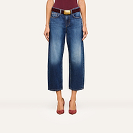 A person wearing the Mallis Bow-Leg Jean in indigo wash, paired with a burgundy belt featuring a large buckle and burgundy pointed-toe heels, stands against a plain background. The upper body is partially visible.