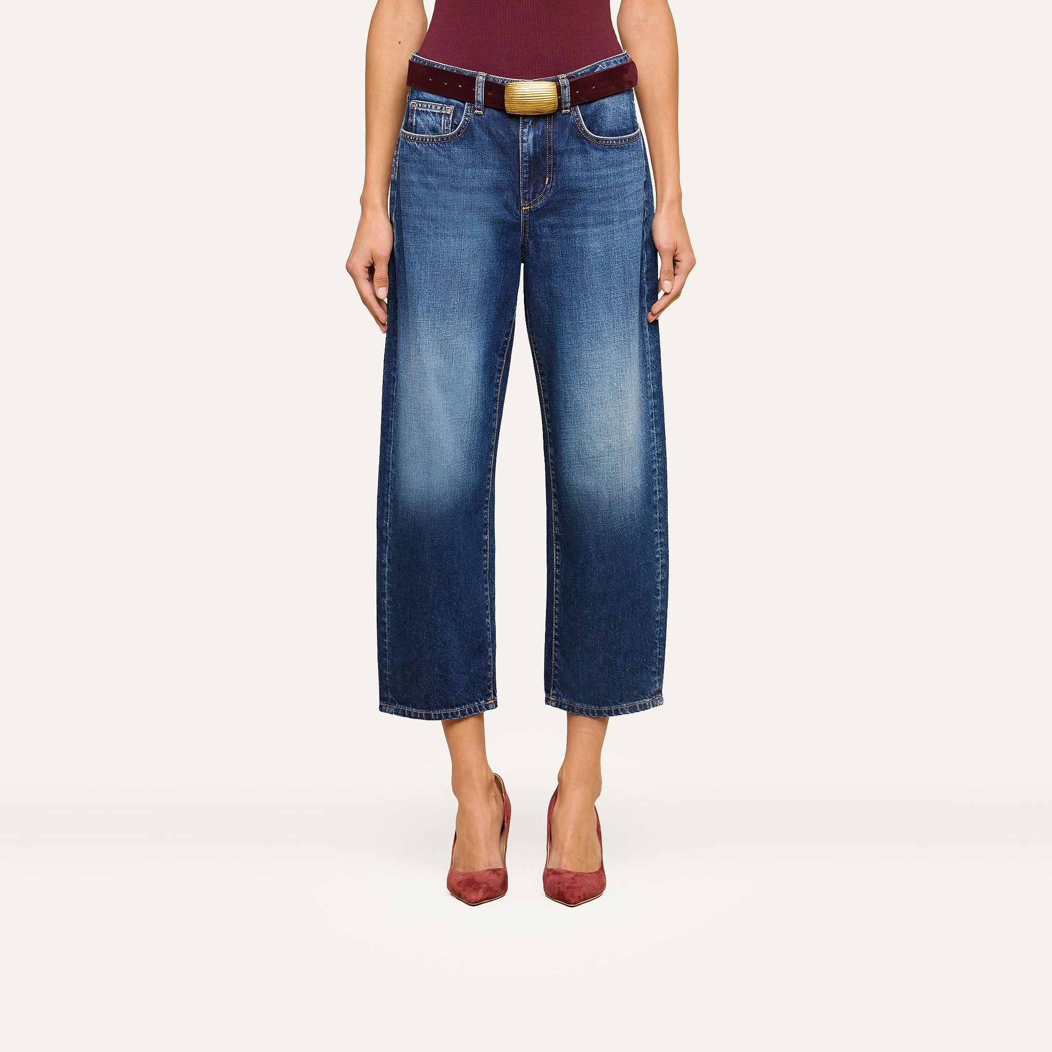 A person wearing the Mallis Bow-Leg Jean in indigo wash, paired with a burgundy belt featuring a large buckle and burgundy pointed-toe heels, stands against a plain background. The upper body is partially visible.