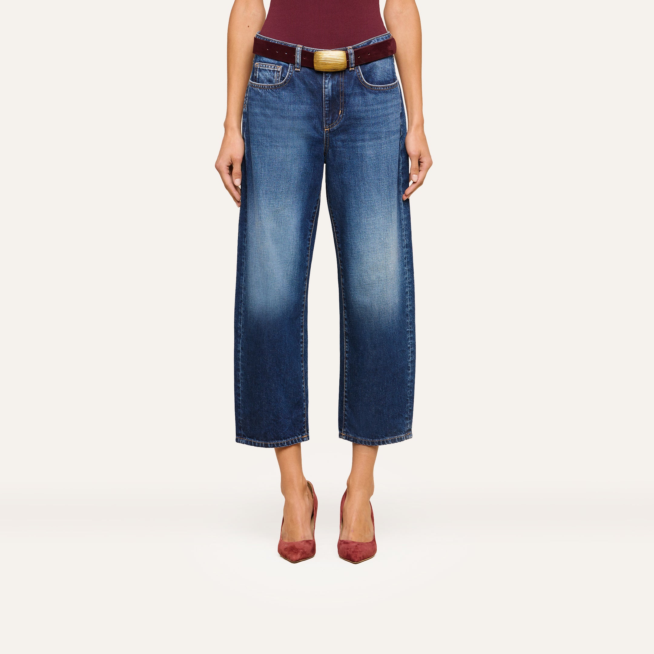 A person wearing the Mallis Bow-Leg Jean in indigo wash, paired with a burgundy belt featuring a large buckle and burgundy pointed-toe heels, stands against a plain background. The upper body is partially visible.