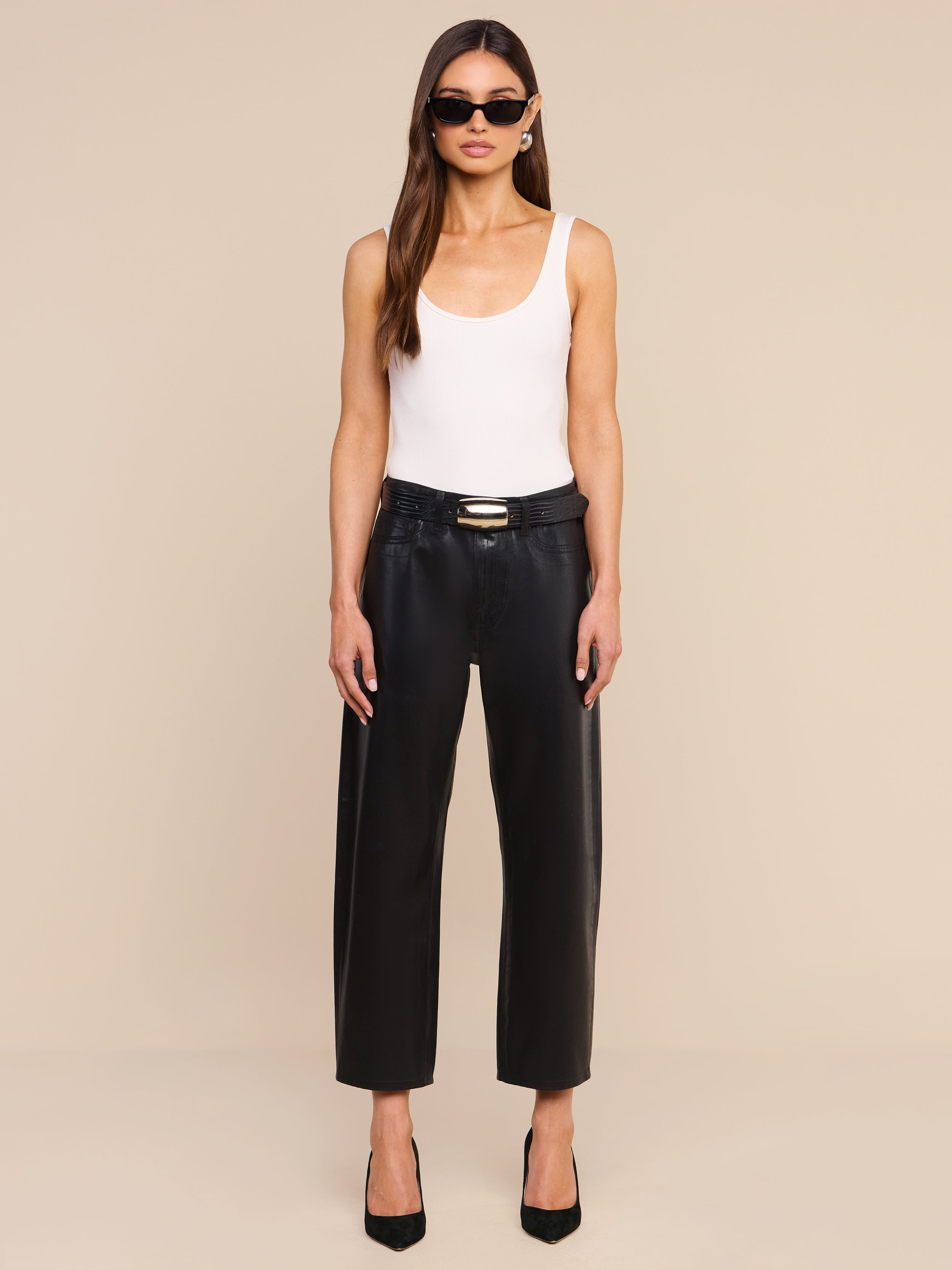 A woman stands against a plain white background, facing forward with her arms relaxed by her sides, wearing sunglasses, a white tank top, black heels, and the Mallis Coated Bow-Leg Jean in a high-rise, wide-leg silhouette.