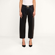 A person wearing the Mallis Coated Bow-Leg Jean—black coated denim pants in a high-rise, wide-leg silhouette—paired with a black belt, a white sleeveless top, and black heels stands against a plain white background. The person’s head is not visible in the image.