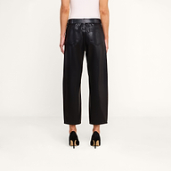 Model standing against a plain background, shown from the back, wearing the Mallis Coated Bow-Leg Jean in black with a high-rise and wide-leg silhouette, paired with black high-heeled shoes and a white sleeveless top.
