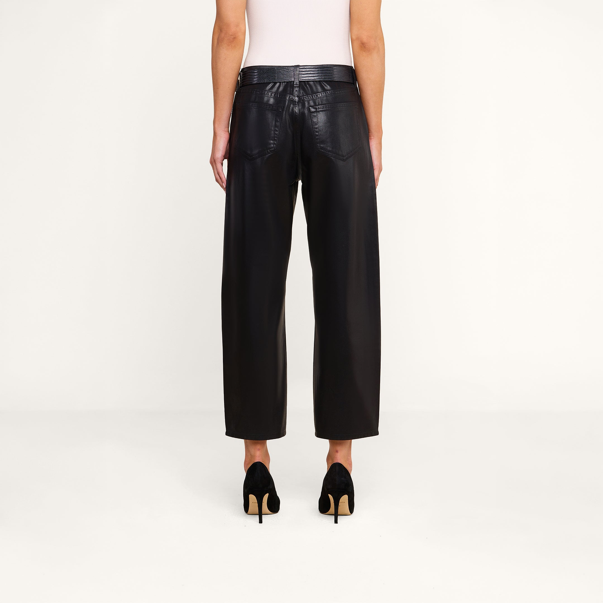Model standing against a plain background, shown from the back, wearing the Mallis Coated Bow-Leg Jean in black with a high-rise and wide-leg silhouette, paired with black high-heeled shoes and a white sleeveless top.