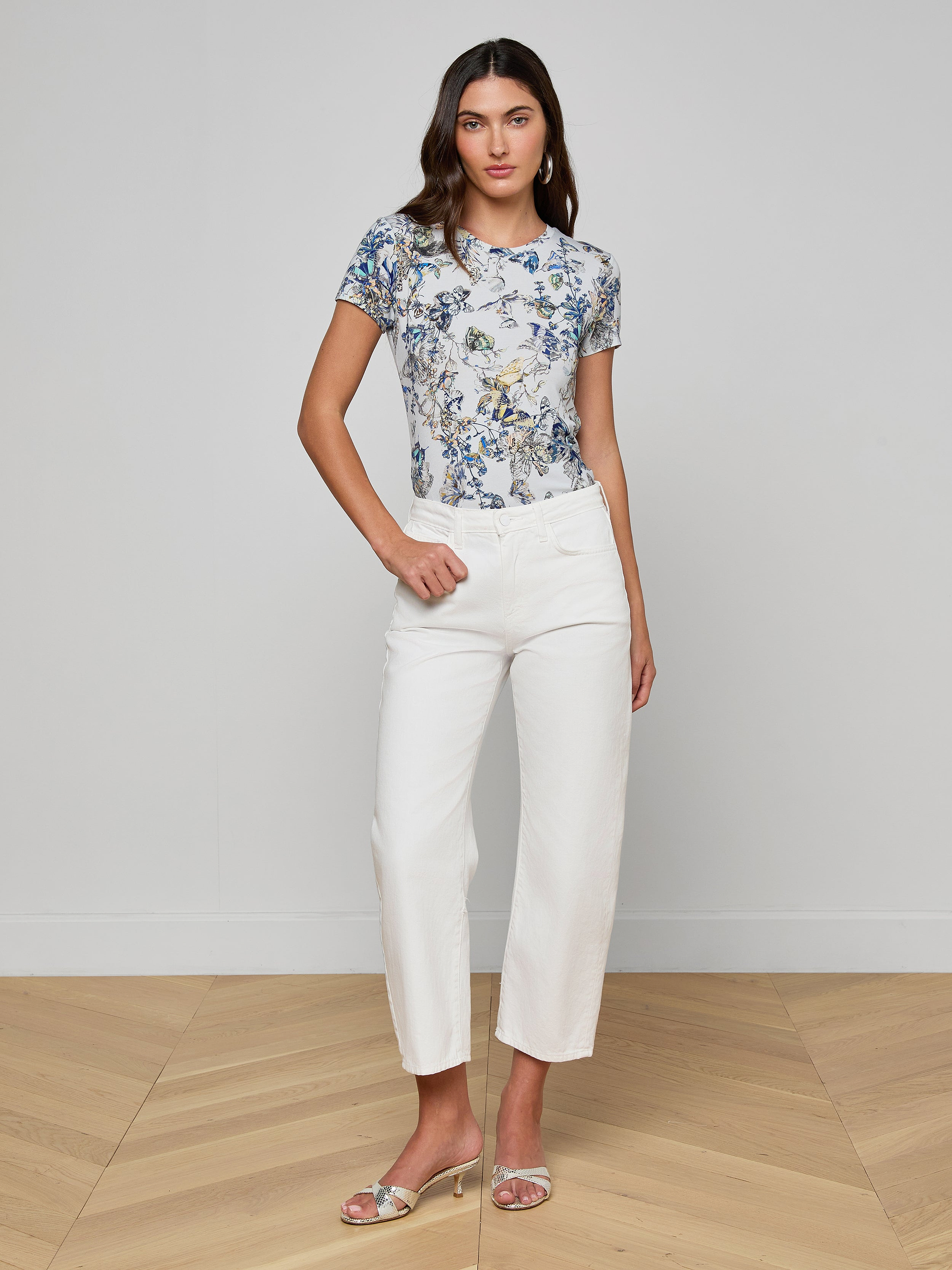 A woman stands against a plain white background, wearing a short-sleeve blue floral top, the Mallis Bow-Leg Jean in a high-rise wide-leg denim style, and open-toe heels. She has one hand on her hip and a neutral expression.