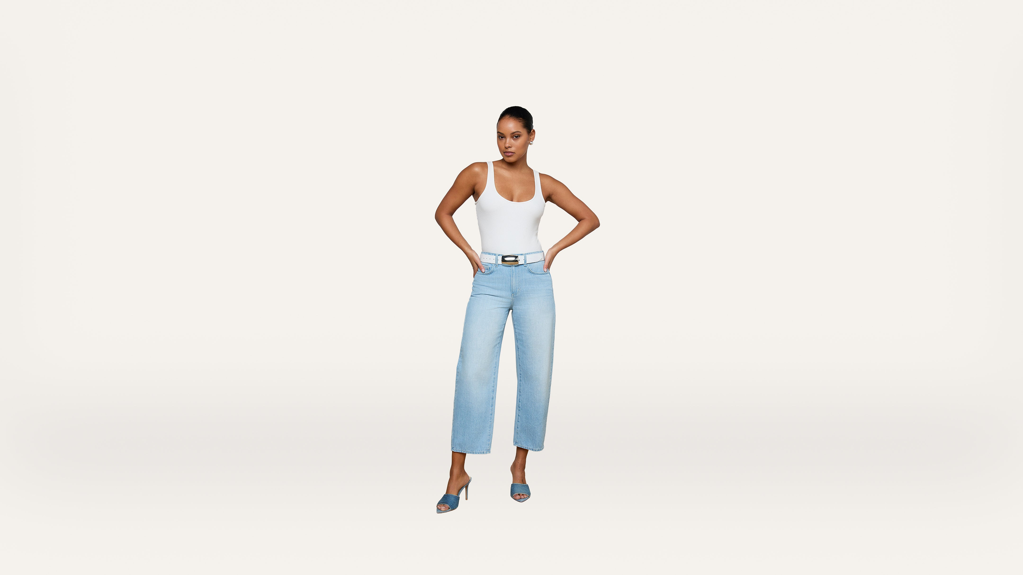A woman stands with hands on hips, wearing a white tank top, light blue high-waisted jeans, and blue heeled sandals against a plain white background.