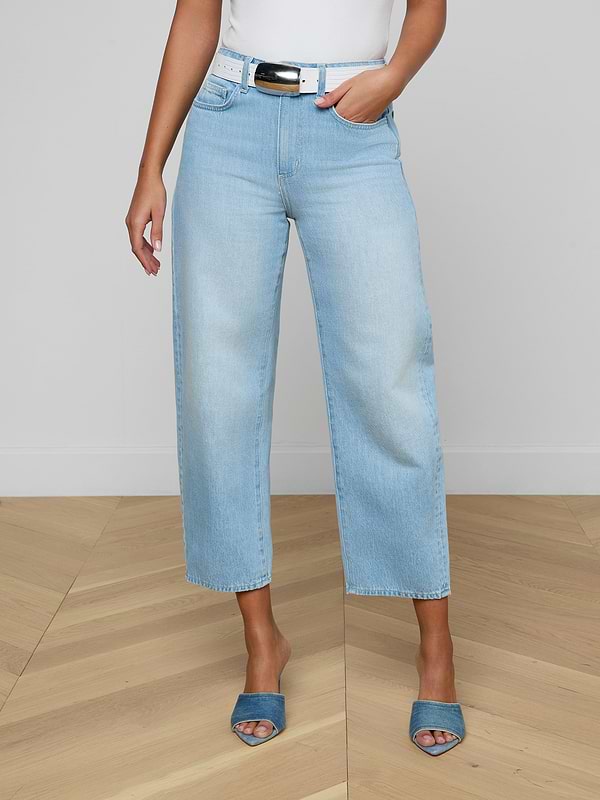 A person stands against a plain white background wearing the Mallis Bow-Leg Jean in light blue with a wide-leg, modern silhouette, paired with a white top and blue open-toe heels. The high-rise denim is styled fashionably, and the image is cropped above the shoulders.