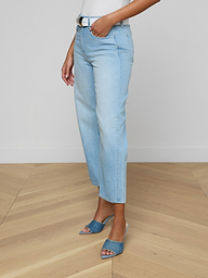 Woman wearing the Mallis Bow-Leg Jean in a light wash with a high rise and straight-leg fit, paired with a white top and blue open-toe heeled sandals, standing against a plain white background. Only the lower half of her body is visible.