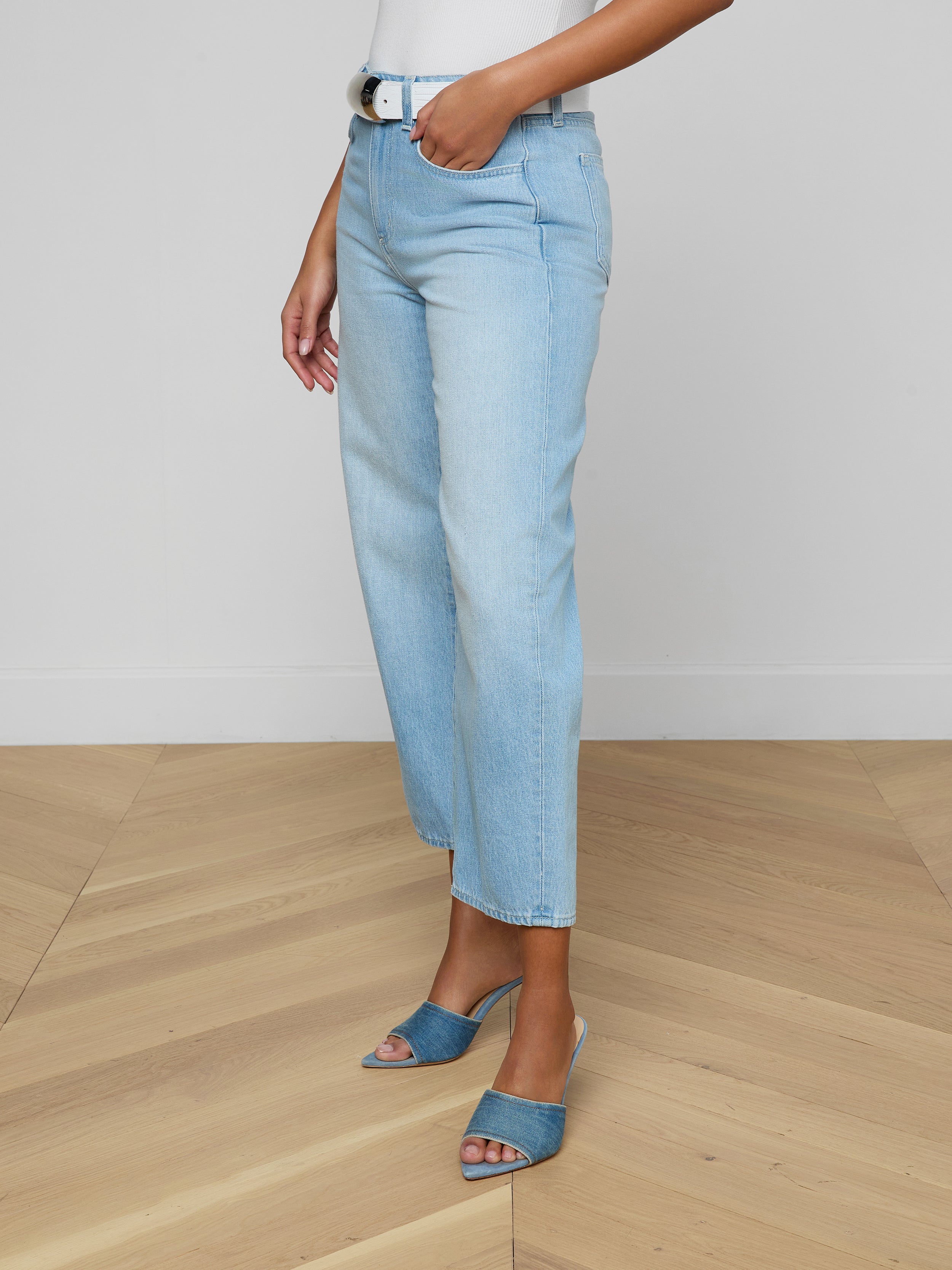 Woman wearing the Mallis Bow-Leg Jean in a light wash with a high rise and straight-leg fit, paired with a white top and blue open-toe heeled sandals, standing against a plain white background. Only the lower half of her body is visible.