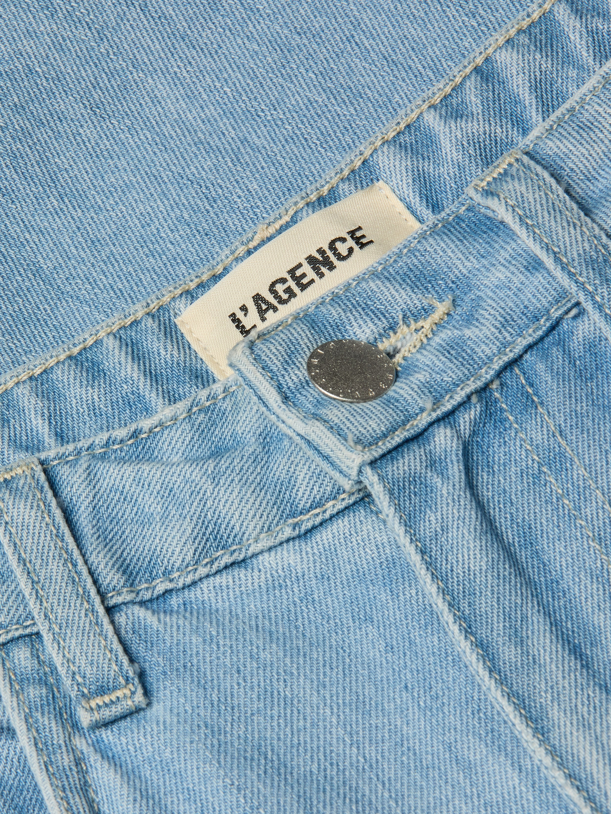 Close-up of the Mallis Bow-Leg Jean in light wash denim, showcasing a silver button and a visible white LAGENCE label with black text inside the waistband.