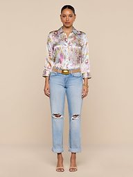 A woman stands facing forward against a plain white background, wearing a vintage-inspired floral button-up shirt, the Ashby Low-Rise Slouch Jean in light blue with ripped details, a belt, and heeled sandals.