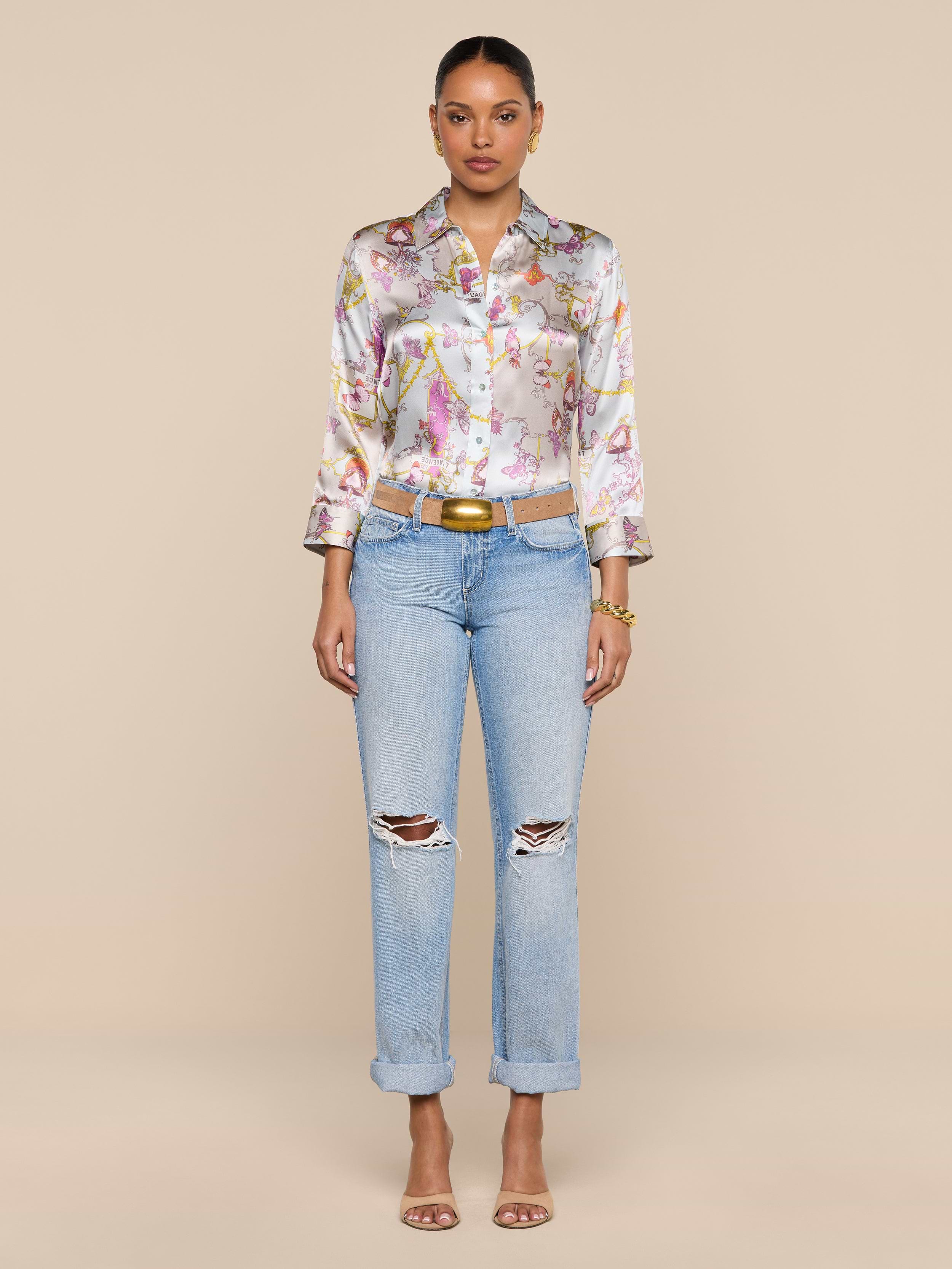 A woman stands facing forward against a plain white background, wearing a vintage-inspired floral button-up shirt, the Ashby Low-Rise Slouch Jean in light blue with ripped details, a belt, and heeled sandals.