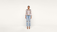 A woman stands facing forward against a plain white background, wearing a floral blouse, ripped light blue jeans, and open-toe heels.