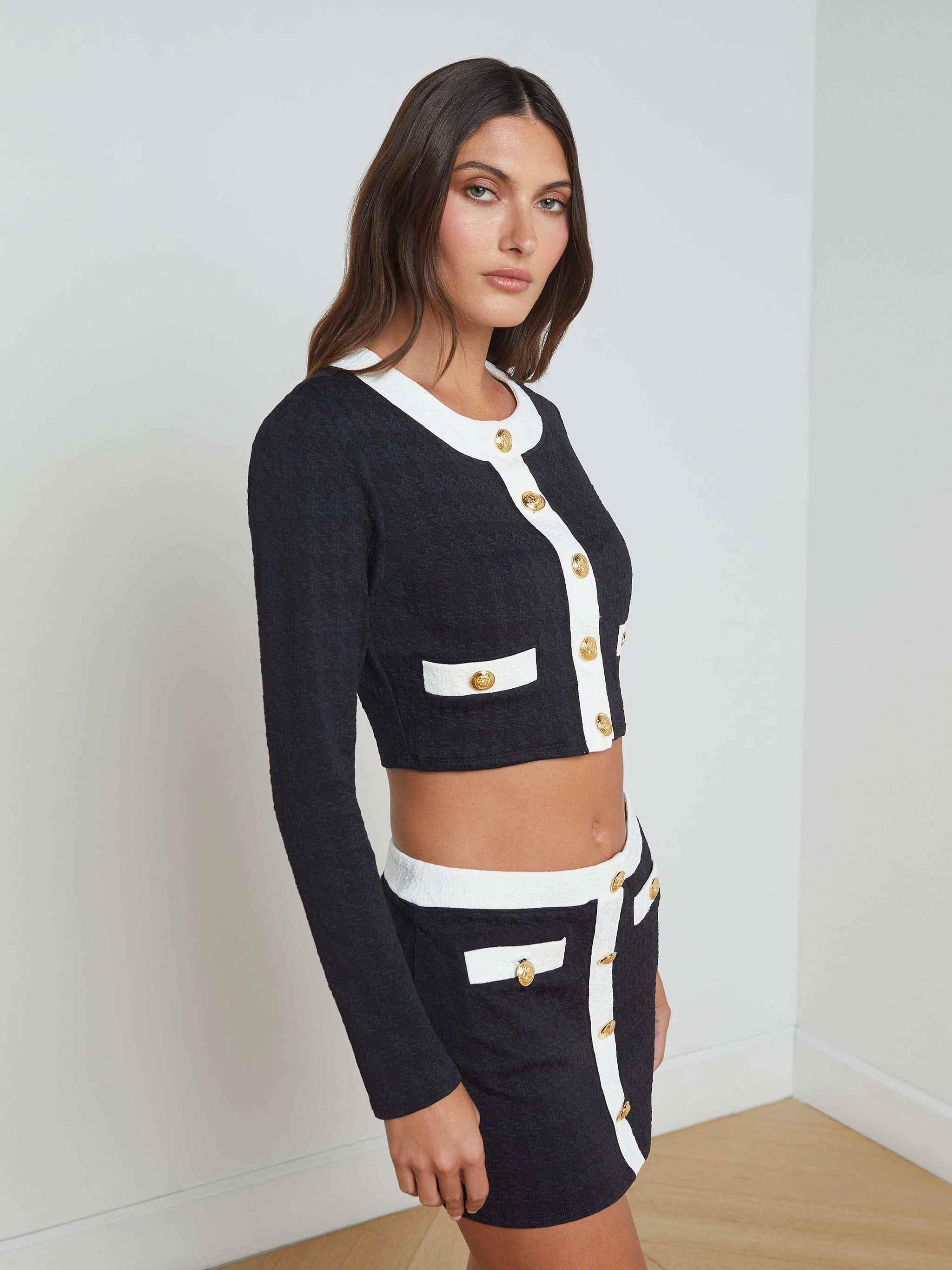 Wearing the Adaline Cardigan Cover-Up—a black and white long-sleeve cropped jacket with gold buttons that’s perfect as a chic swimsuit cover-up—paired with a high-waisted mini skirt, she poses against a plain light background.