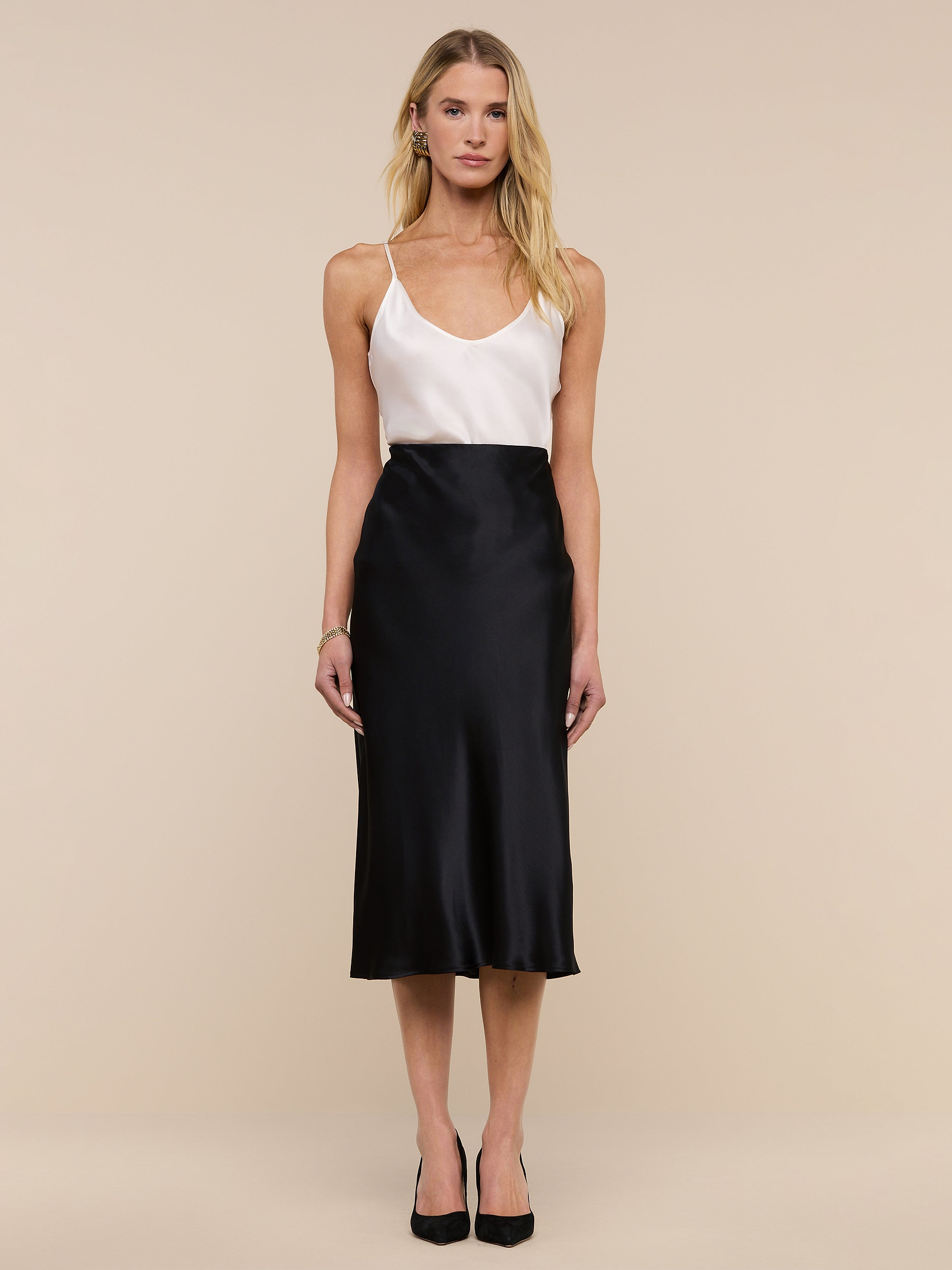 A woman stands against a plain background, wearing a white sleeveless top, the Perin Silk Midi Skirt in black, and black heels. She has long blonde hair and a neutral expression.
