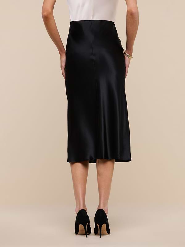 A woman stands facing away against a plain white background, wearing a white sleeveless top, the Perin Silk Midi Skirt in black, and black high-heeled shoes.