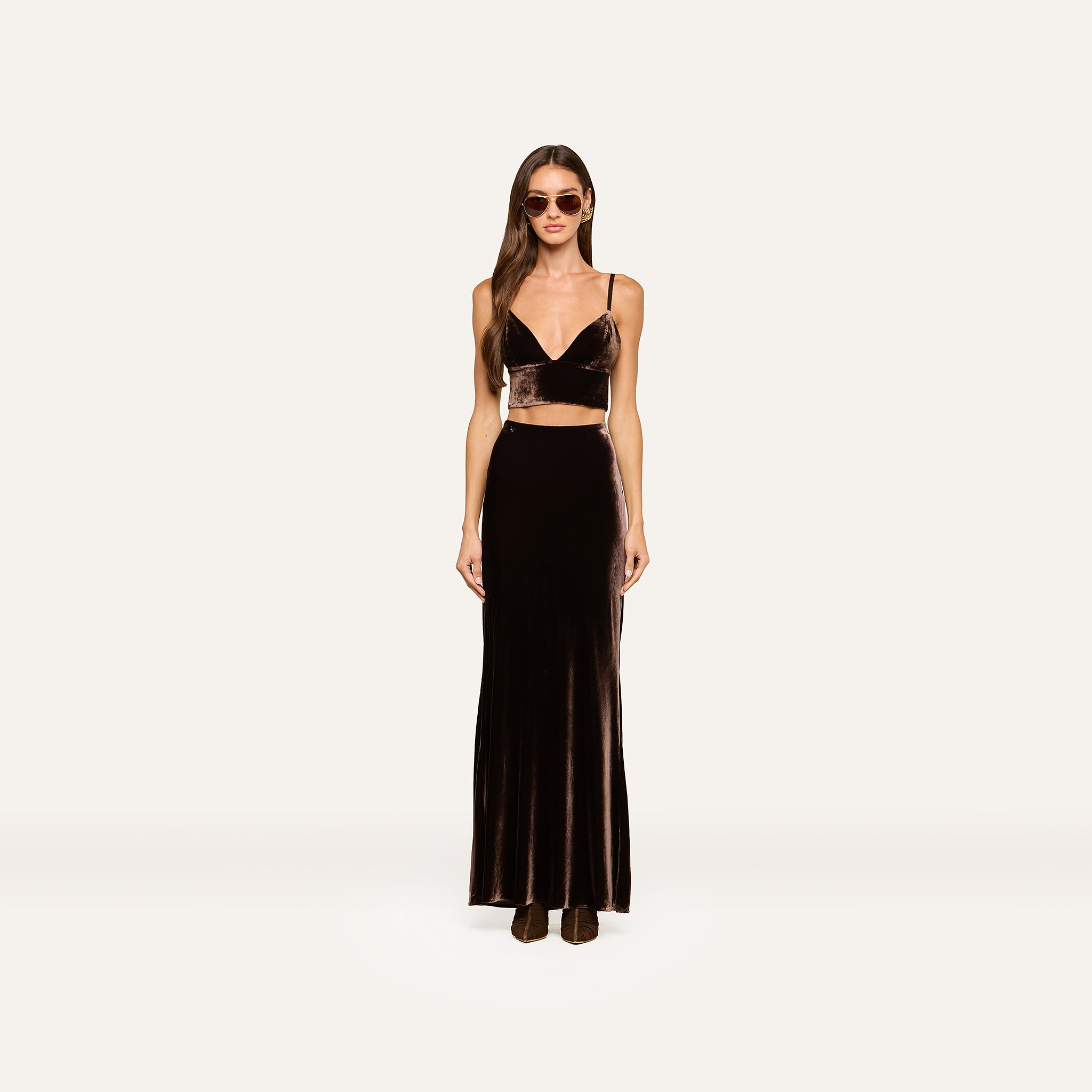 A woman wearing sunglasses stands facing forward in a dark brown velvet crop top and the Zeta Velvet Maxi Skirt with boots, against a plain light background, exemplifying a minimalist wardrobe.