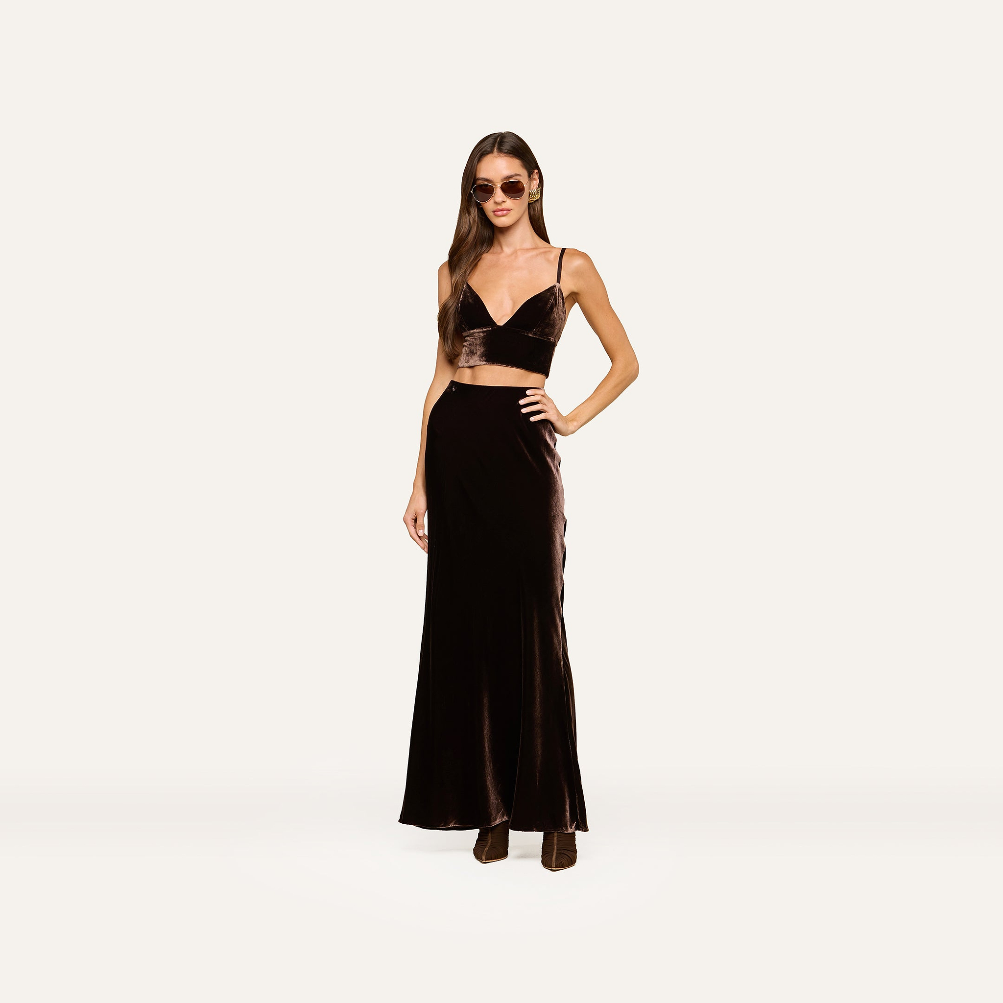A woman wearing sunglasses and a chic black velvet two-piece outfit, featuring a crop top and the Zeta Velvet Maxi Skirt, stands against a plain, light background—exemplifying minimalist style with her sophisticated maxi skirt ensemble.