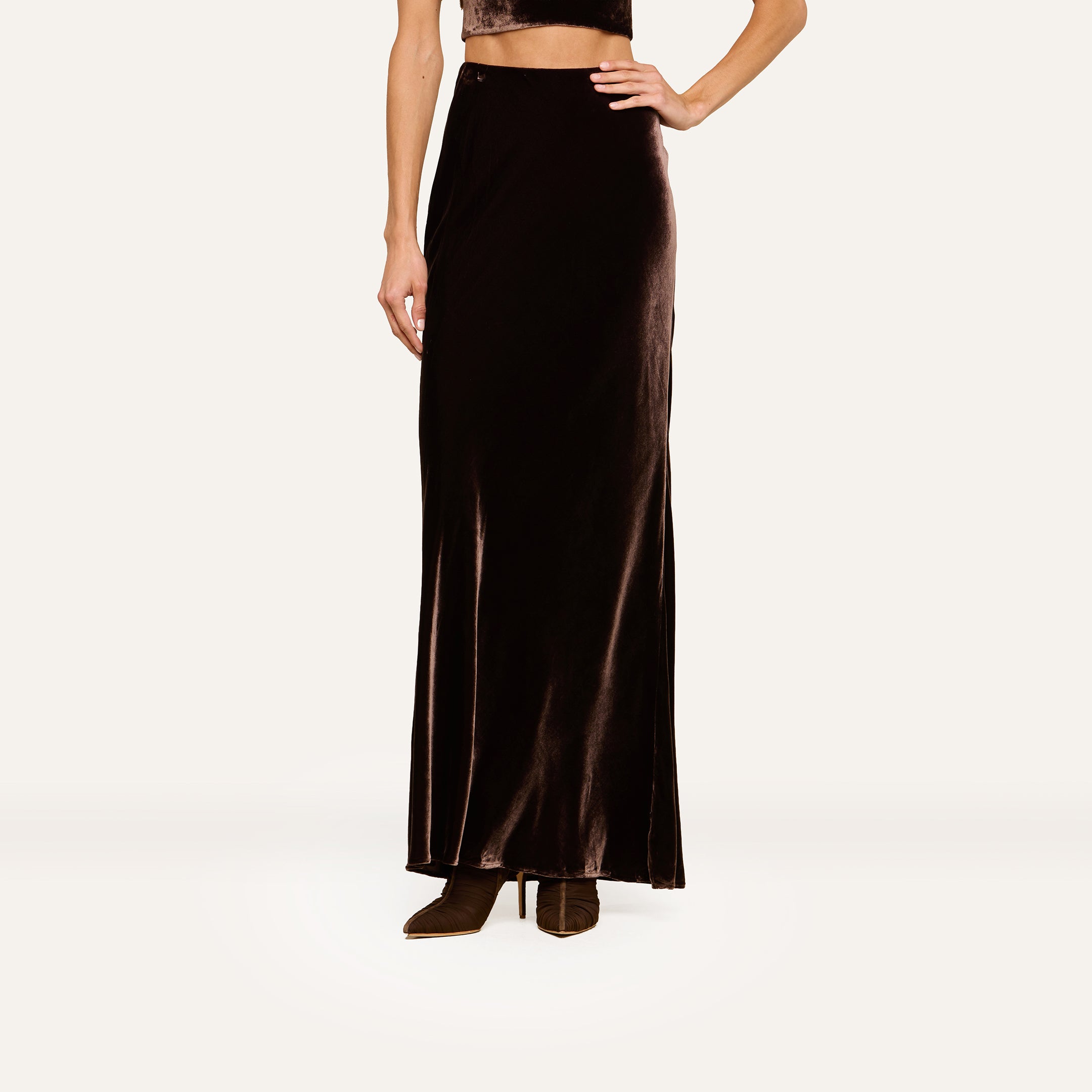 A person models the Zeta Velvet Maxi Skirt with brown boots and a cropped top, standing against a plain white background. The image focuses on the lower half of the body, highlighting a minimalist wardrobe style.