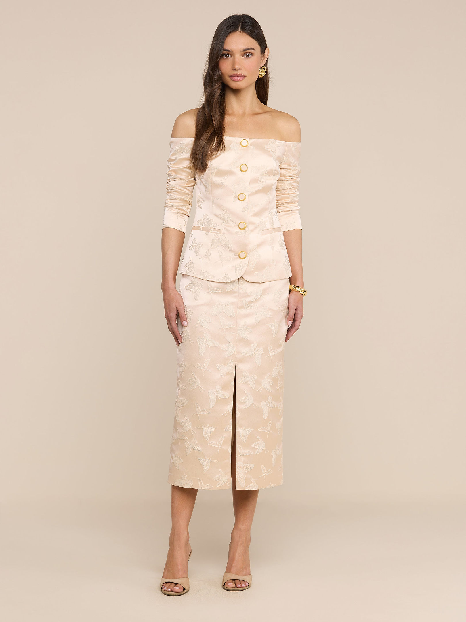 A woman stands against a plain white background wearing the Alice Blazer Top, a cream-colored, off-the-shoulder blazer with gold buttons, paired with a matching below-the-knee skirt featuring a front slit and heels.