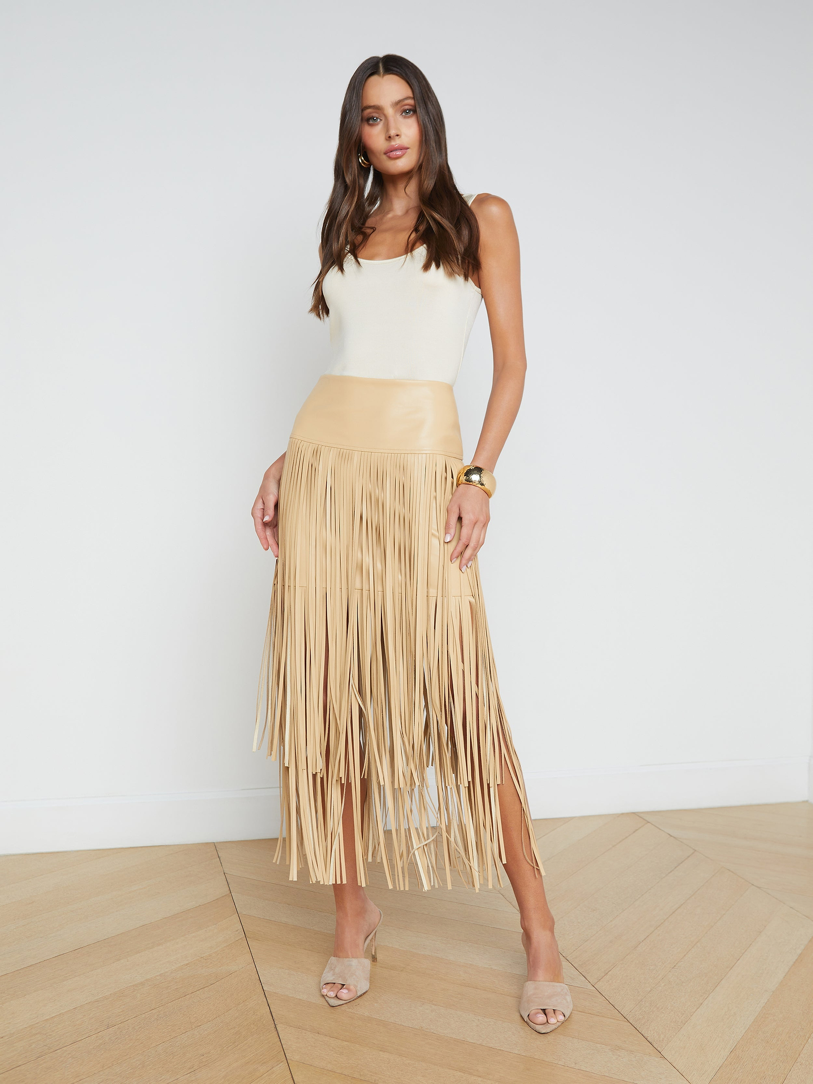 A woman stands facing forward in high heels against a plain white background, wearing a white sleeveless top and the tan Karolina Fringe Skirt made from vegan leather.