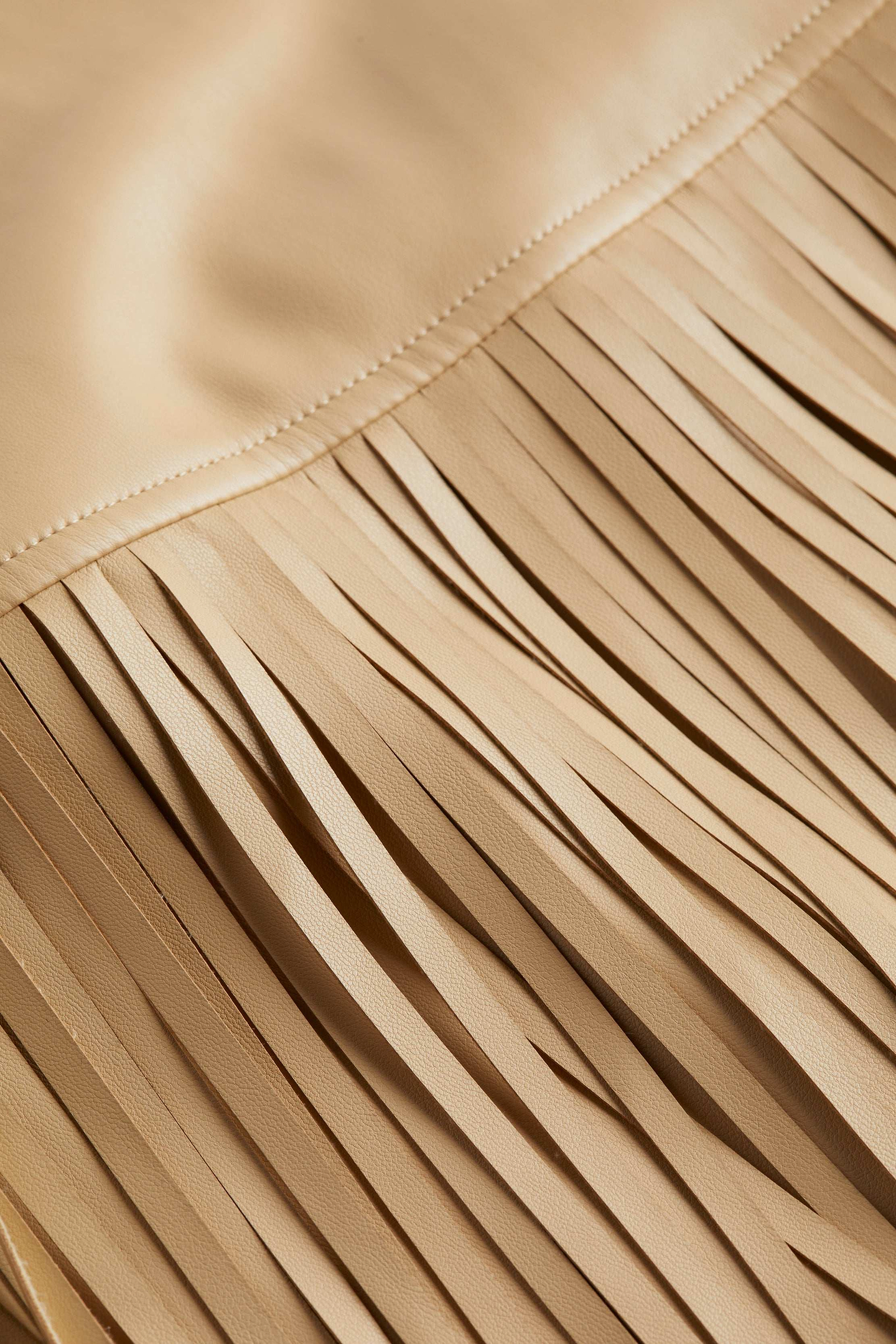 Close-up of beige vegan leather fabric from the Karolina Fringe Skirt, highlighting a stitched seam and long, narrow fringes cascading down for a textured, layered effect reminiscent of a statement piece.