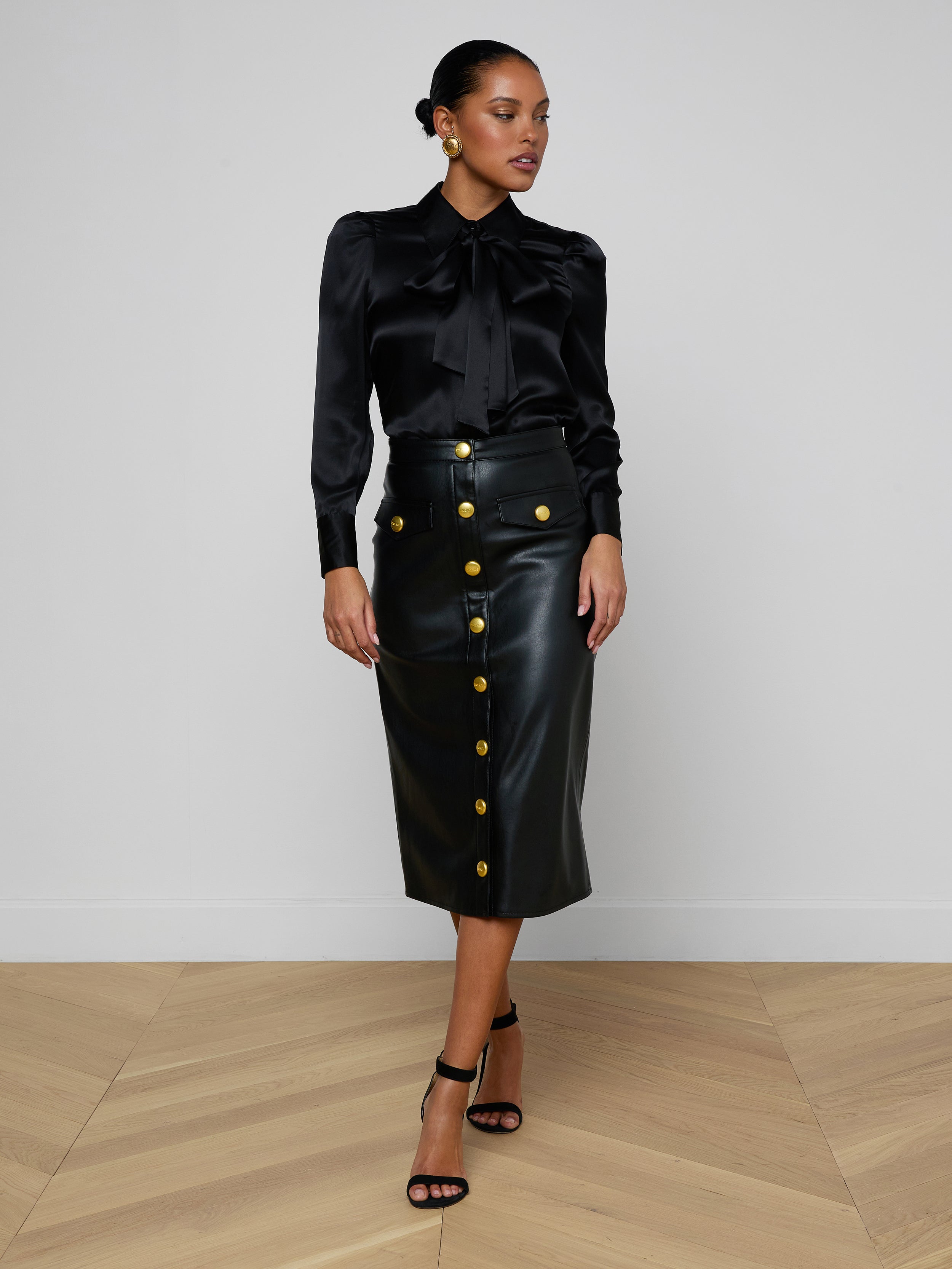 A woman stands confidently against a plain light background, wearing a black satin blouse paired with the high-waisted Milann Vegan Leather Skirt, which features gold buttons, and finishes the look with black heeled sandals.