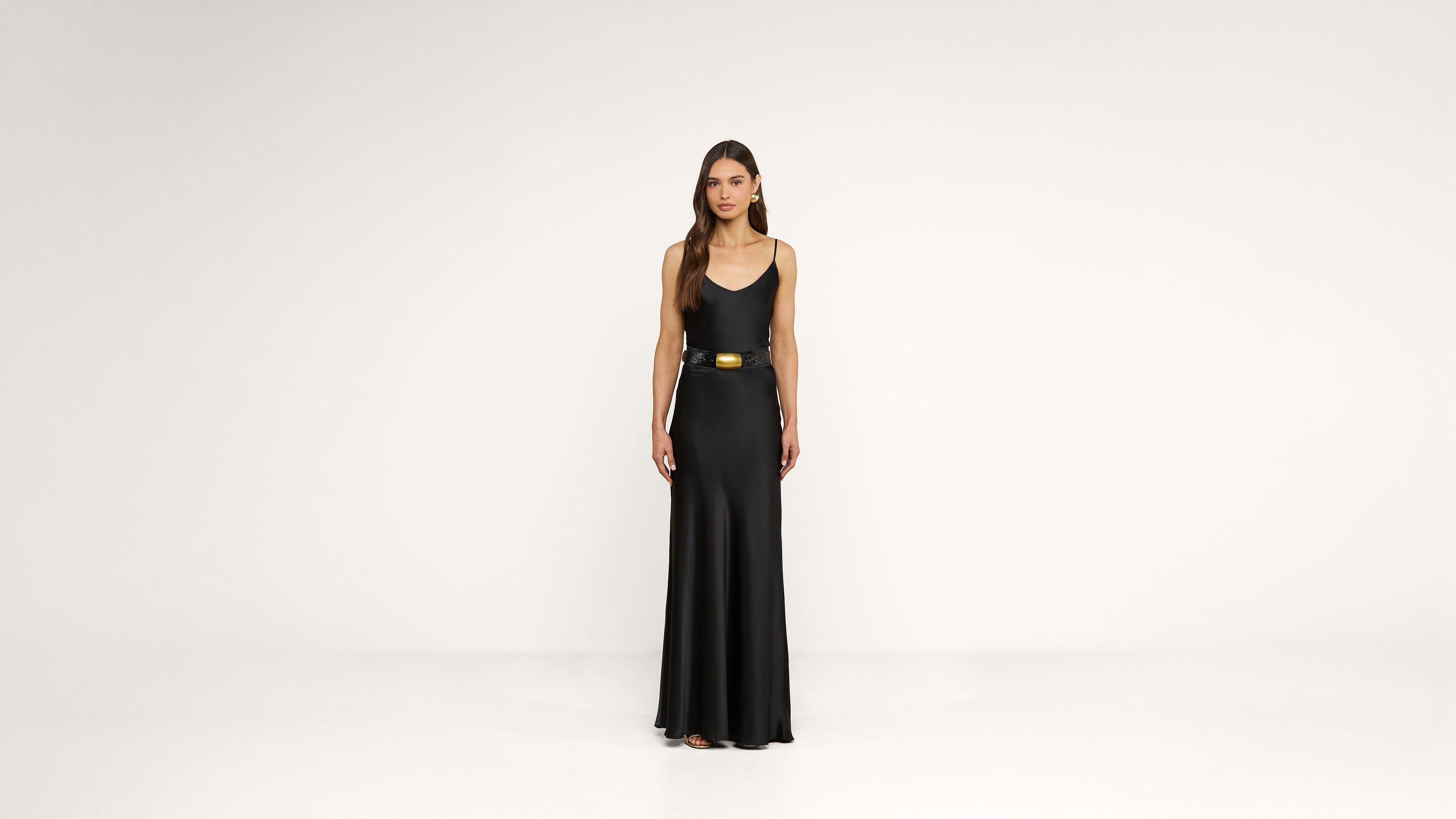 A woman stands facing forward, wearing a long, sleeveless black dress with a gold belt, against a plain white background.