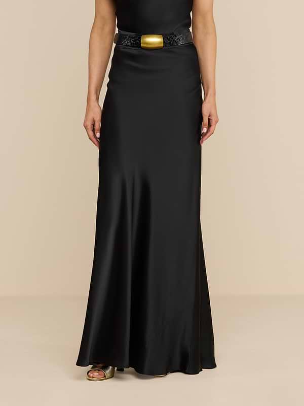 A person wearing the Ryker Silk Maxi Skirt, a long black bias-cut skirt, paired with a black belt featuring a large gold buckle, standing against a plain white background.