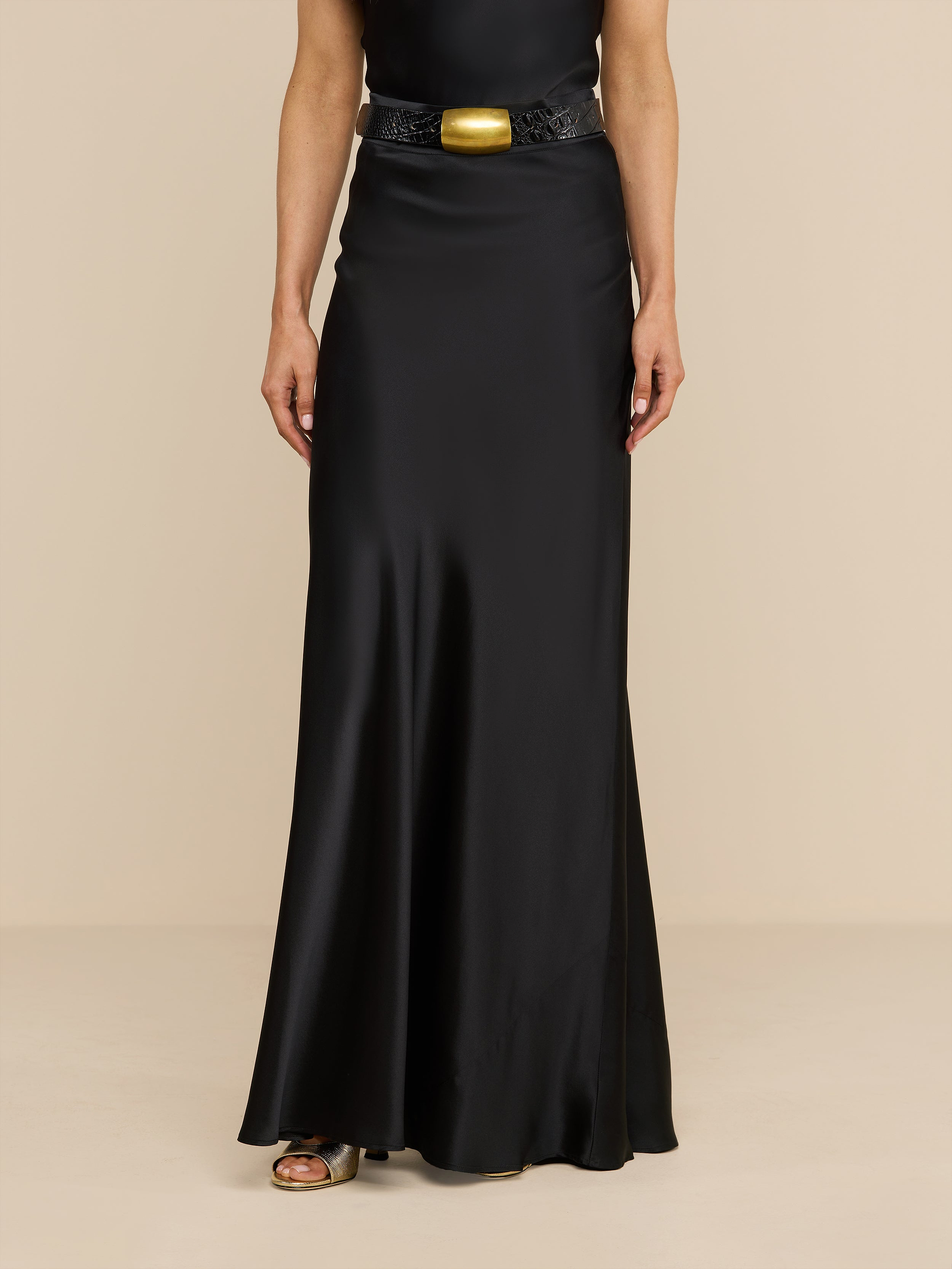 A person wearing the Ryker Silk Maxi Skirt, a long black bias-cut skirt, paired with a black belt featuring a large gold buckle, standing against a plain white background.