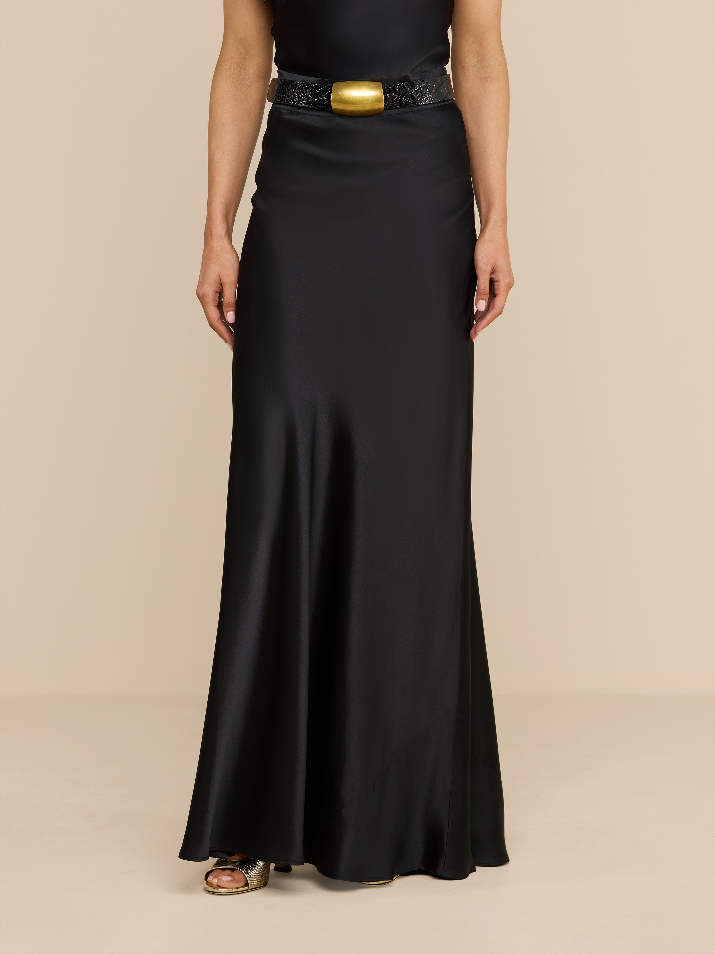 A person wearing the Ryker Silk Maxi Skirt, a long black bias-cut skirt, paired with a black belt featuring a large gold buckle, standing against a plain white background.