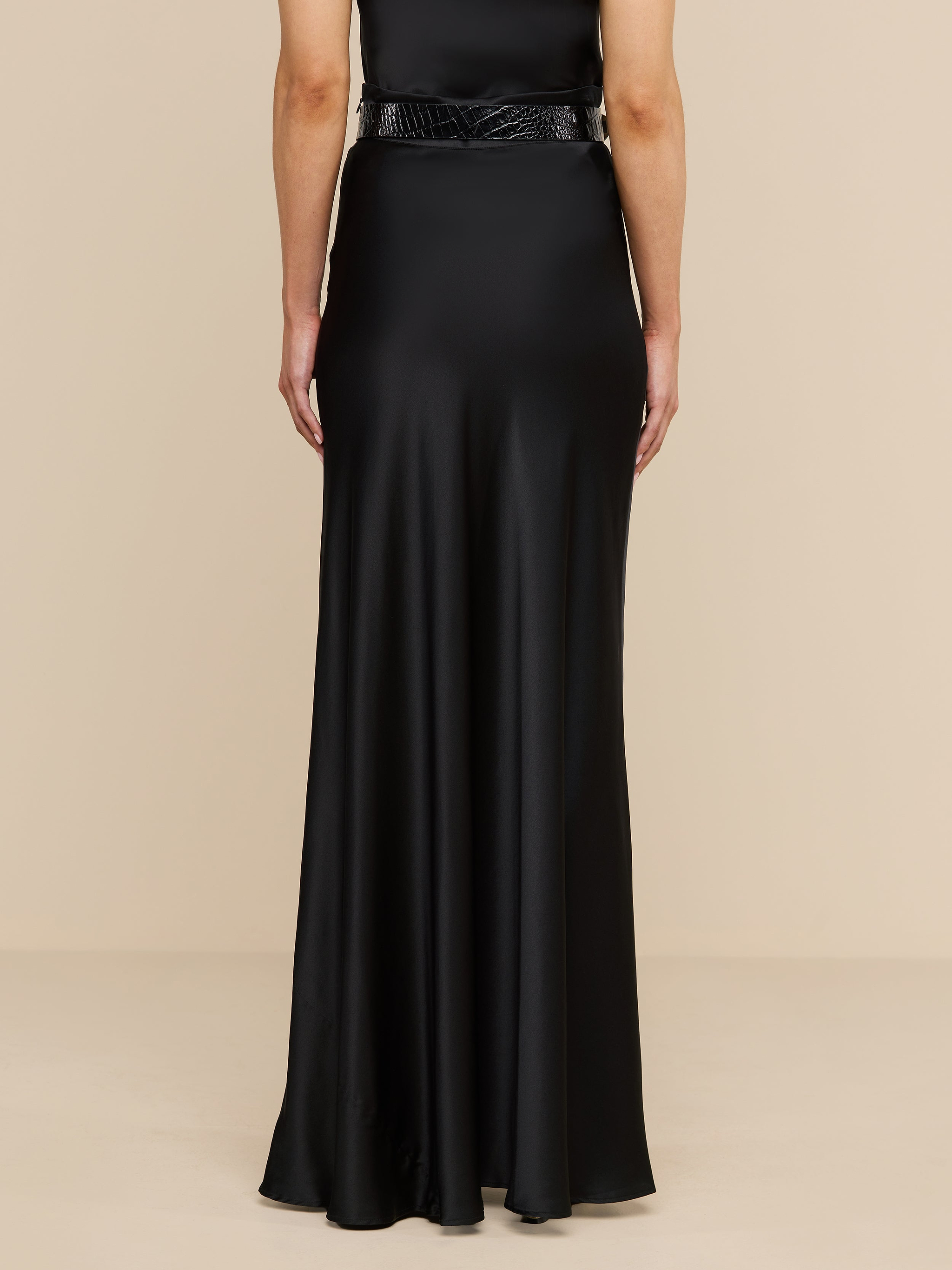 A person stands facing away, wearing the Ryker Silk Maxi Skirt in black, paired with a minimalist black belt for an effortlessly sophisticated look. The background is plain and white.