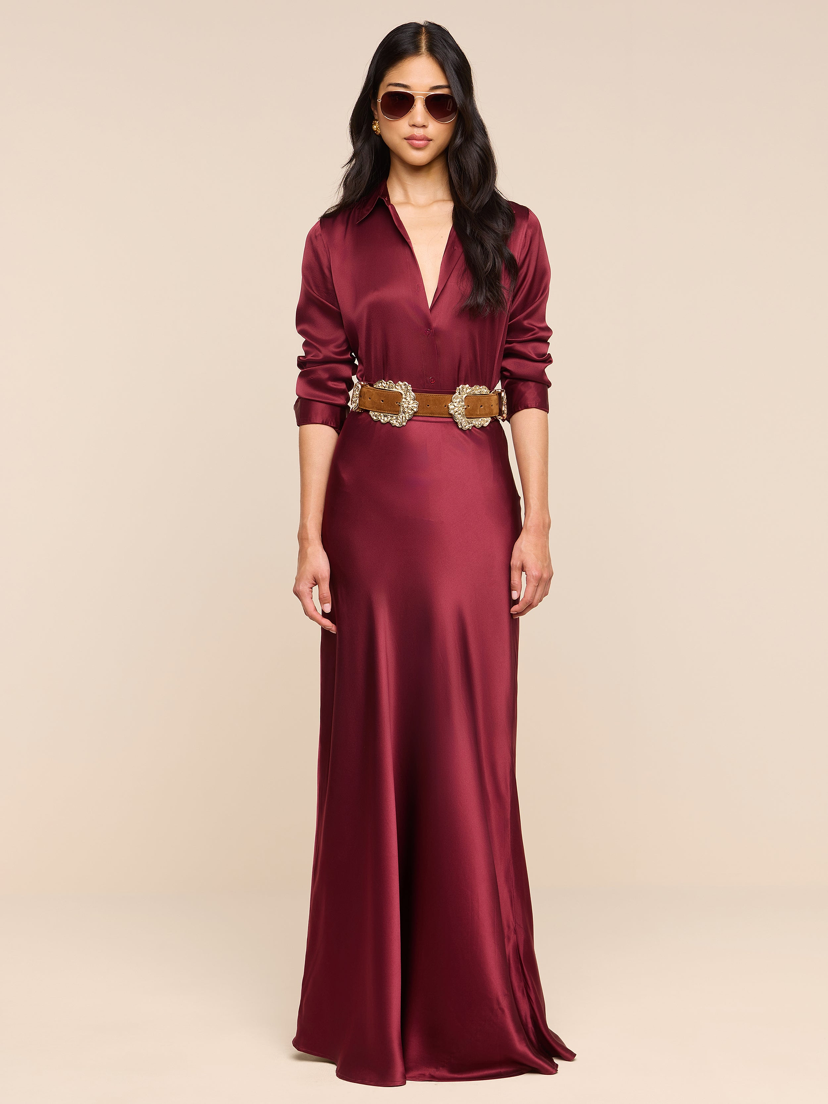 A woman stands against a plain white background, wearing the Ryker Silk Maxi Skirt in merlot red paired with a brown belt and sunglasses, looking forward with her hands by her sides.