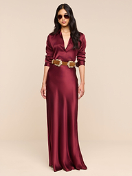 A woman stands confidently against a plain white background wearing the Ryker Silk Maxi Skirt in merlot red, styled with a brown belt. The skirt features a flattering bias cut. She has long dark hair, sunglasses, and her hands are relaxed by her sides.
