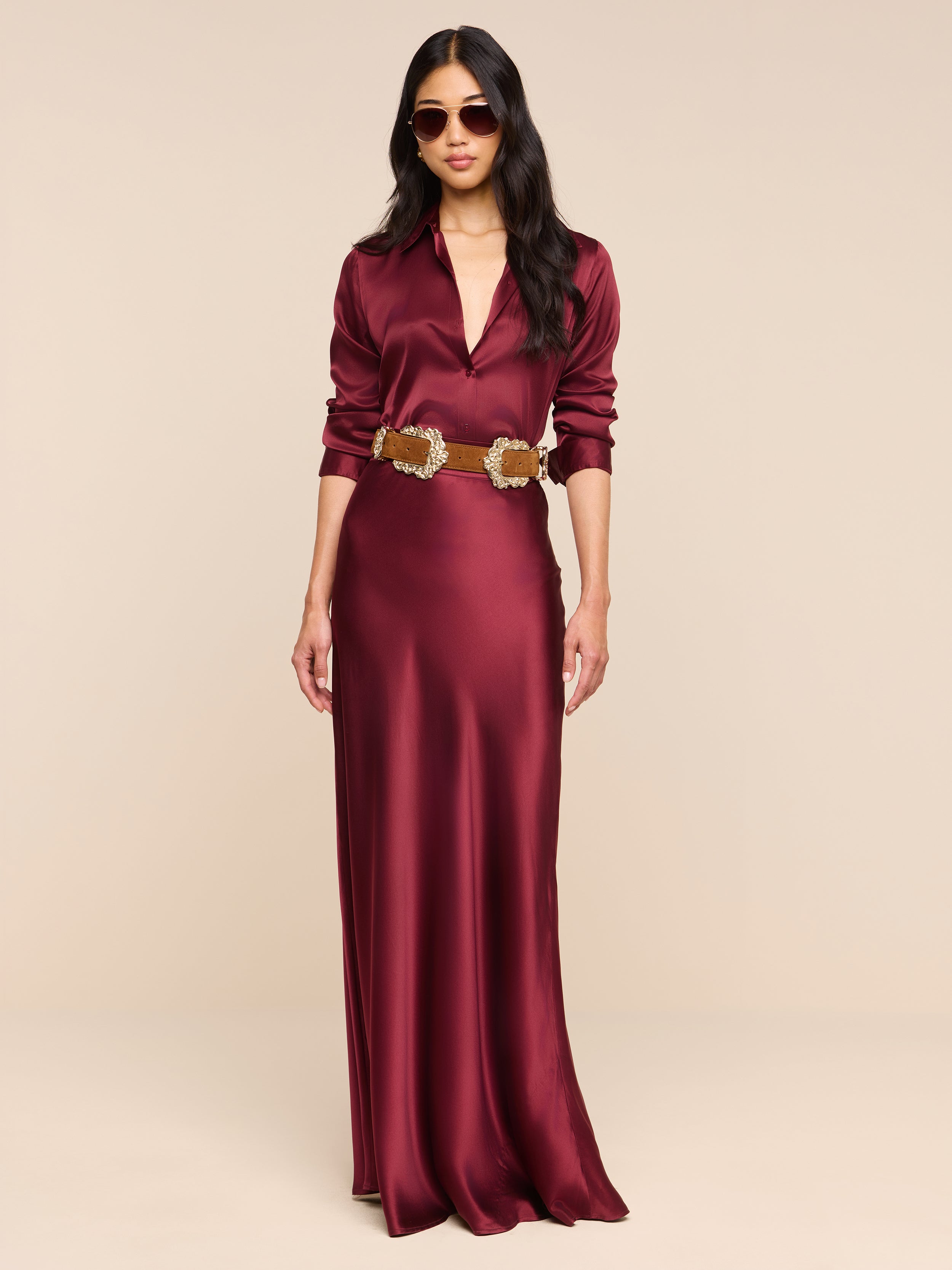 A woman stands confidently against a plain white background wearing the Ryker Silk Maxi Skirt in merlot red, styled with a brown belt. The skirt features a flattering bias cut. She has long dark hair, sunglasses, and her hands are relaxed by her sides.
