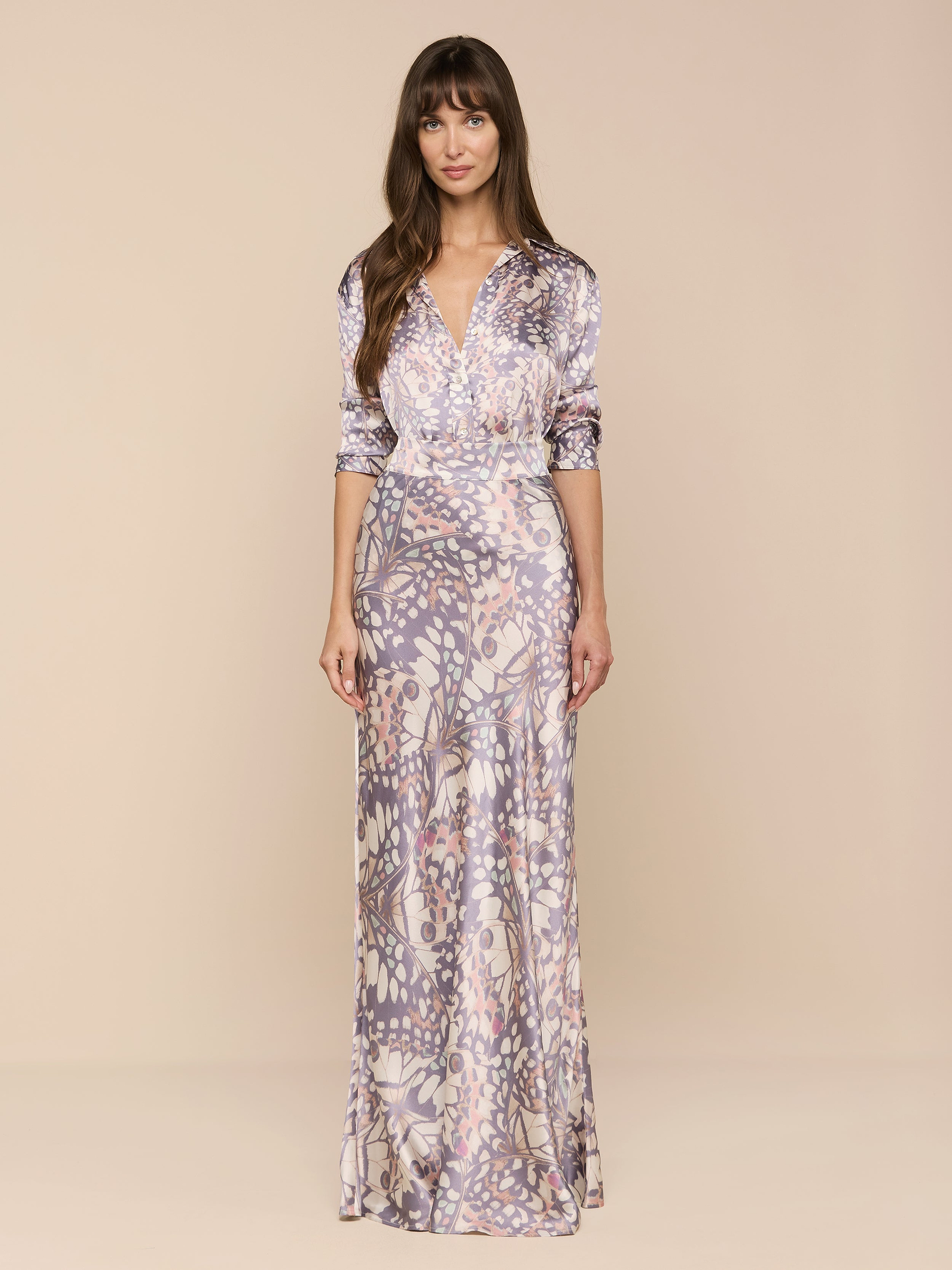 Una donna dai lunghi capelli castani e dallespressione rilassata si staglia su uno sfondo beige, indossando la maxi gonna Ryker Silk con stampa di ali di farfalla color pastello, abbinata a un top a maniche lunghe coordinato con scollo a V.