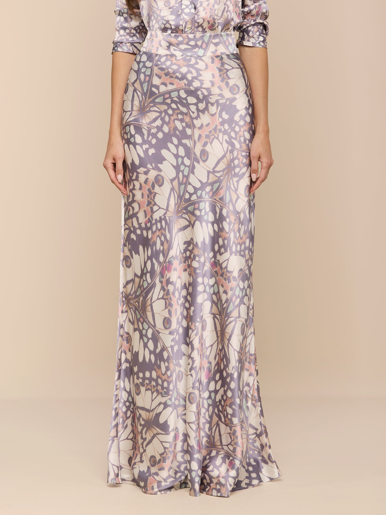 Una donna indossa la maxi gonna in seta Ryker, caratterizzata da una silhouette lunga e aderente con un motivo botanico e astratto color pastello ispirato alle ali delle farfalle. La texture setosa risalta su uno sfondo beige. La testa della donna non è visibile nellimmagine.