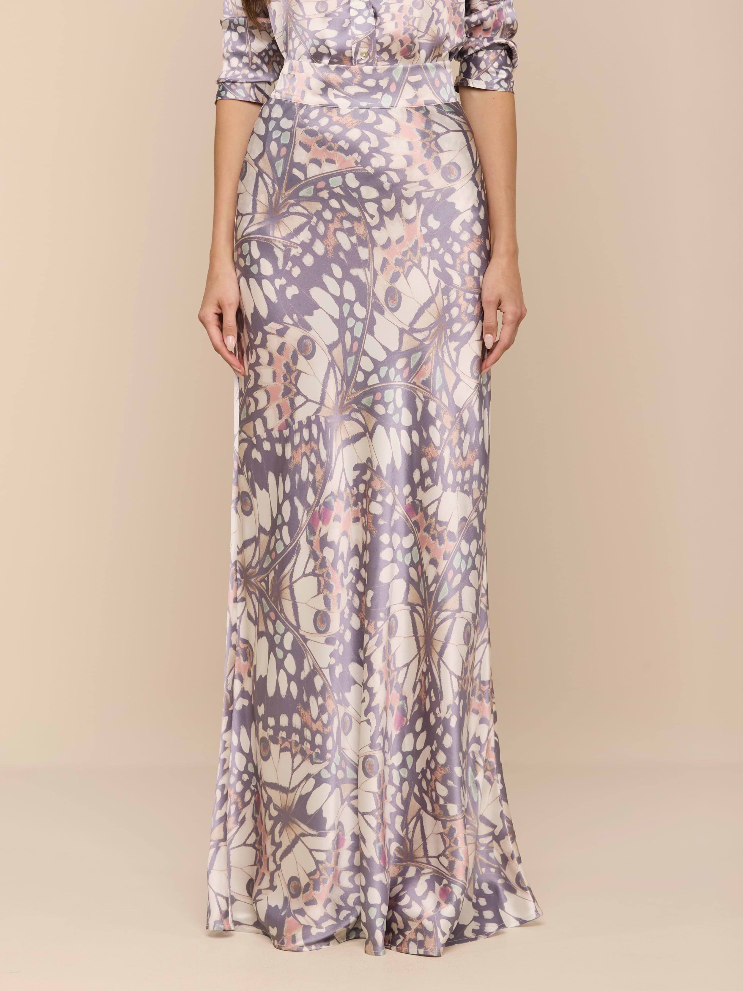 A person stands against a plain white background wearing the Ryker Silk Maxi Skirt in a long, silky, lavender and cream butterfly wing print.