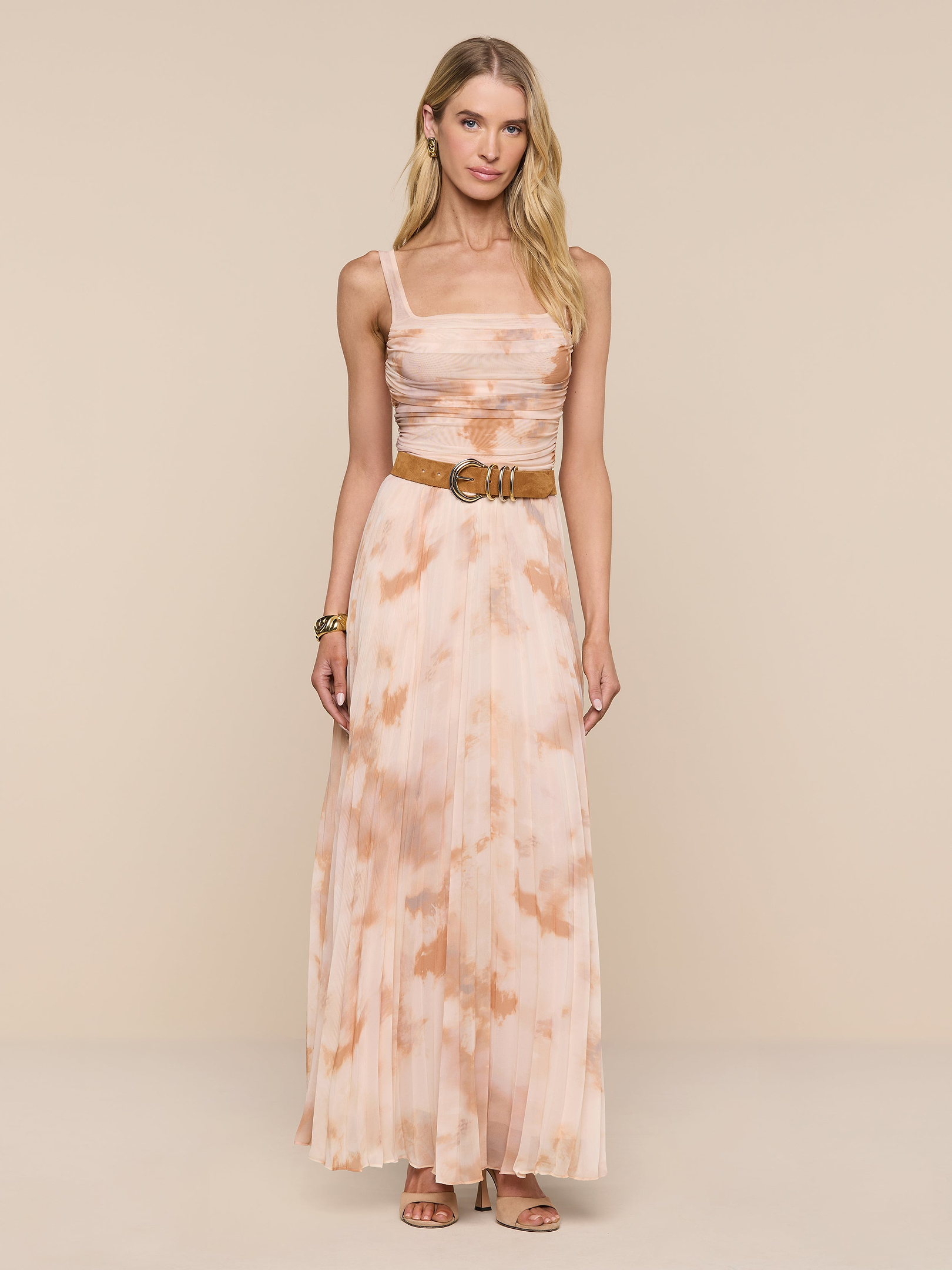 A woman stands against a plain white background wearing the Torin Pleated Maxi Skirt in peach and cream, styled with a wide belt and sandals. She has long blonde hair and accessorizes with bracelets.
