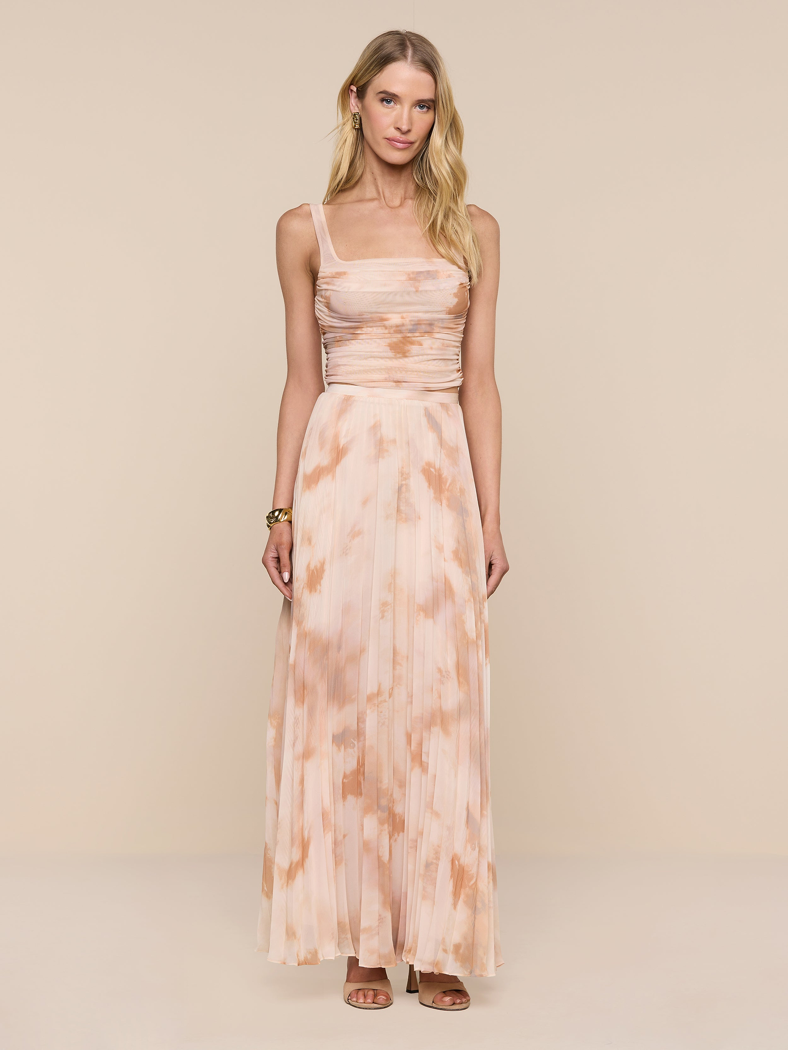 A woman stands against a plain white background, wearing the Torin Pleated Maxi Skirt in a long, peach and beige tie-dye design. She pairs the airy poly chiffon skirt with a sleeveless top featuring thin straps and a ruched bodice. Her long blonde hair flows down, and she accessorizes with brown sandals and a bracelet.