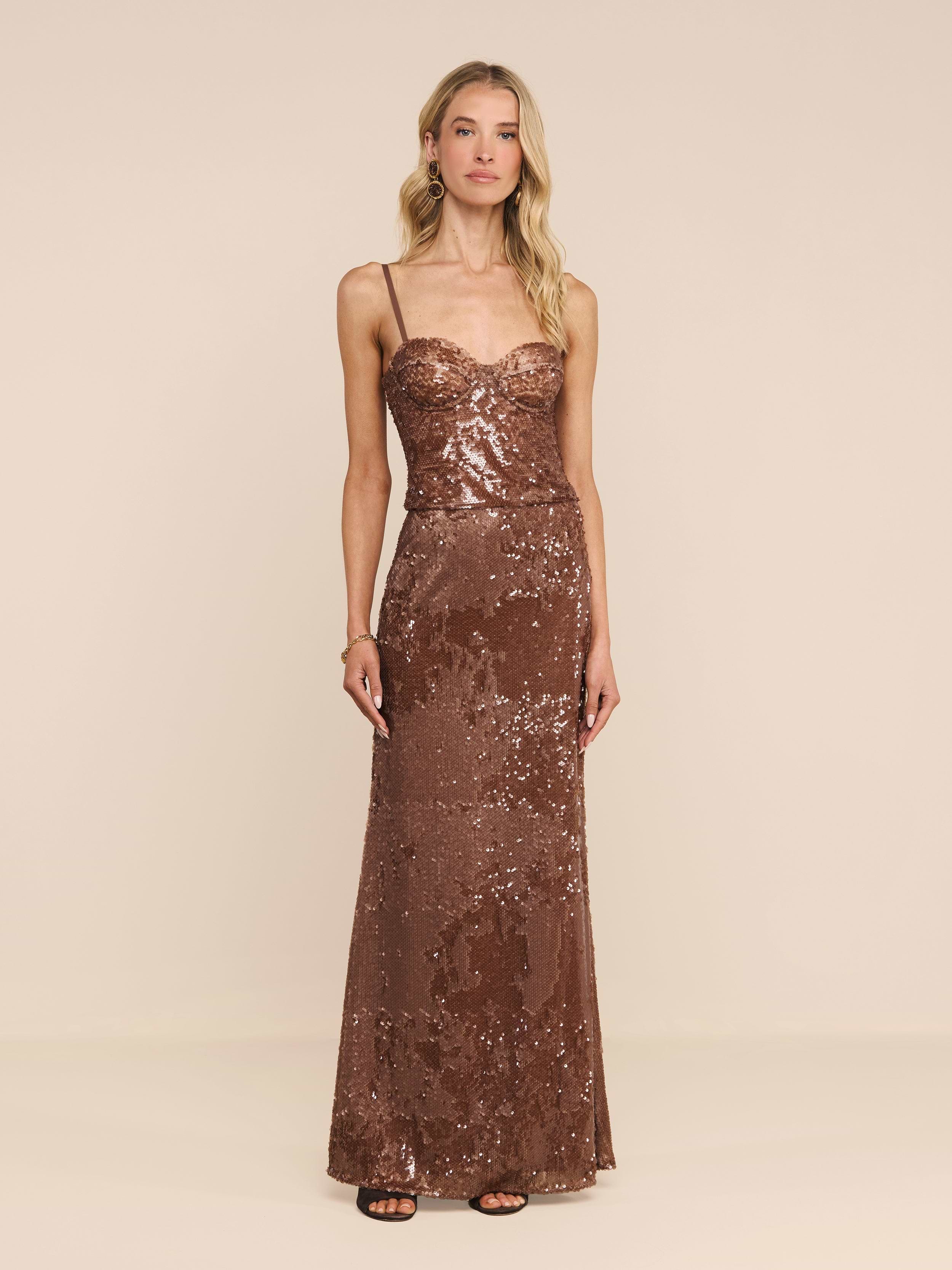 Una donna è in piedi su uno sfondo beige a tinta unita e guarda davanti a sé con espressione neutra, indossando la gonna lunga Zeta Sequin Maxi Skirt, un modello aderente color bronzo che arriva fino a terra, impreziosito da paillettes.